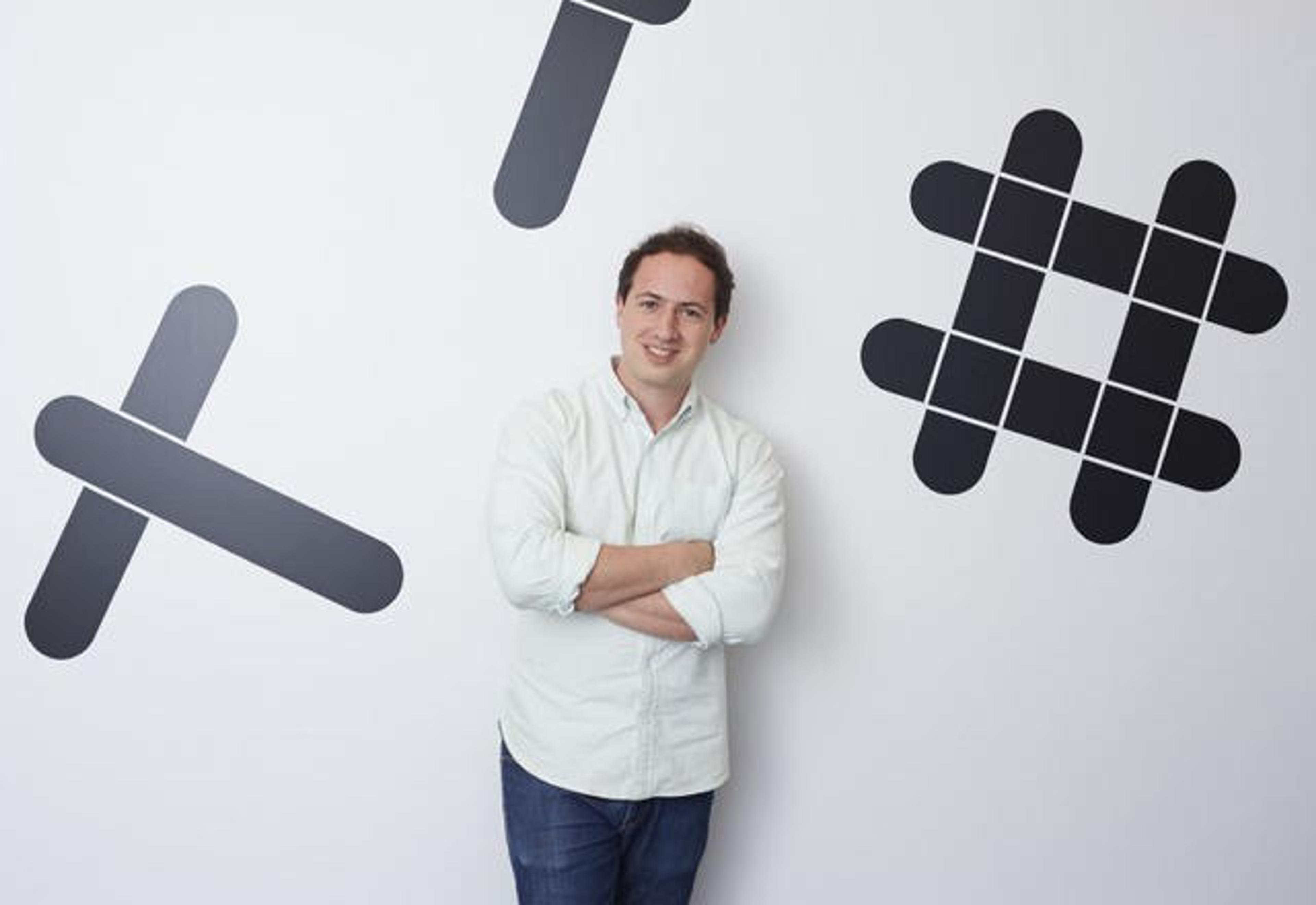 A person stands with arms crossed in front of a wall featuring large black graphic symbols. They are smiling and wearing a light-colored shirt.