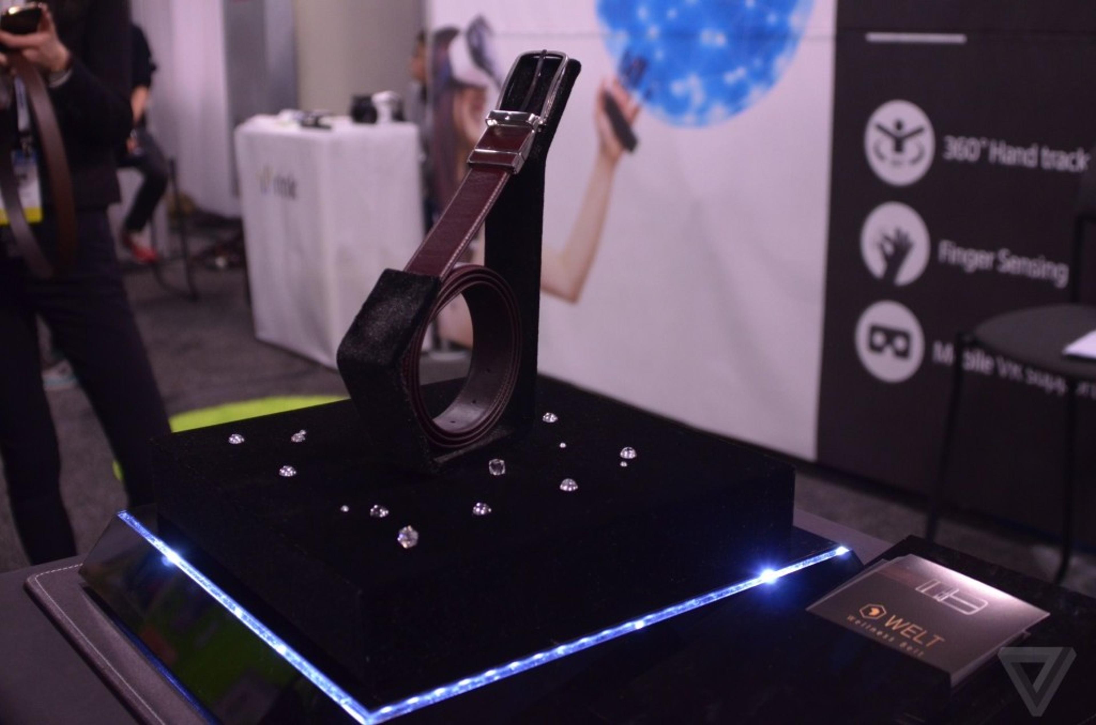 A smart belt displayed on a pedestal with a black velvet surface, surrounded by small lights. The background features a blurred booth with promotional materials.
