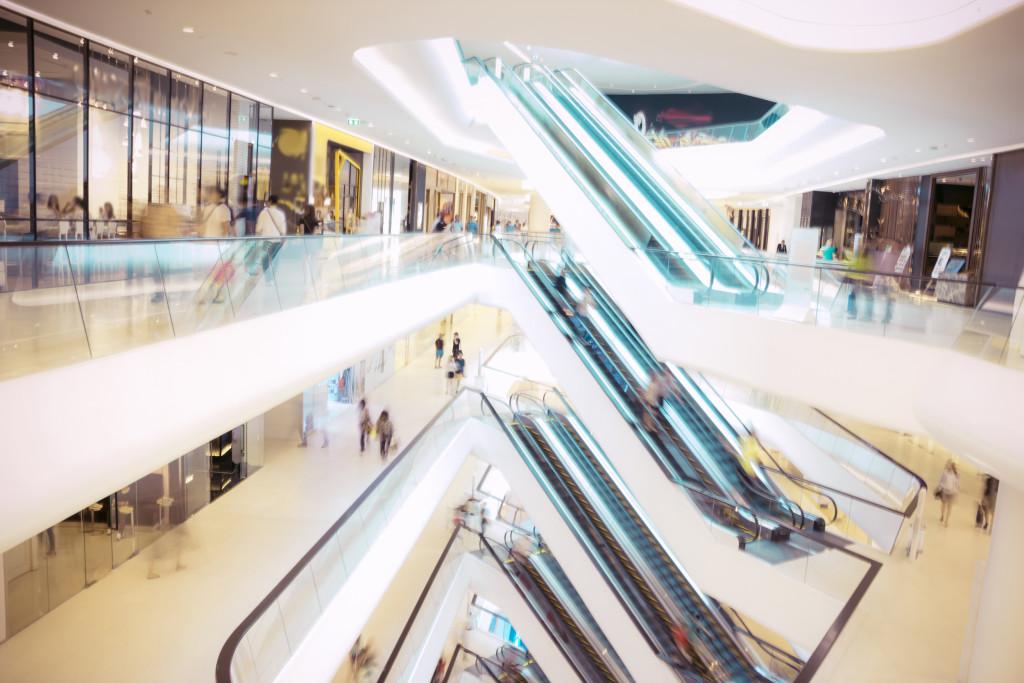 A modern shopping mall interior featuring multiple escalators and a bright, open design. Shoppers are seen moving throughout the space.