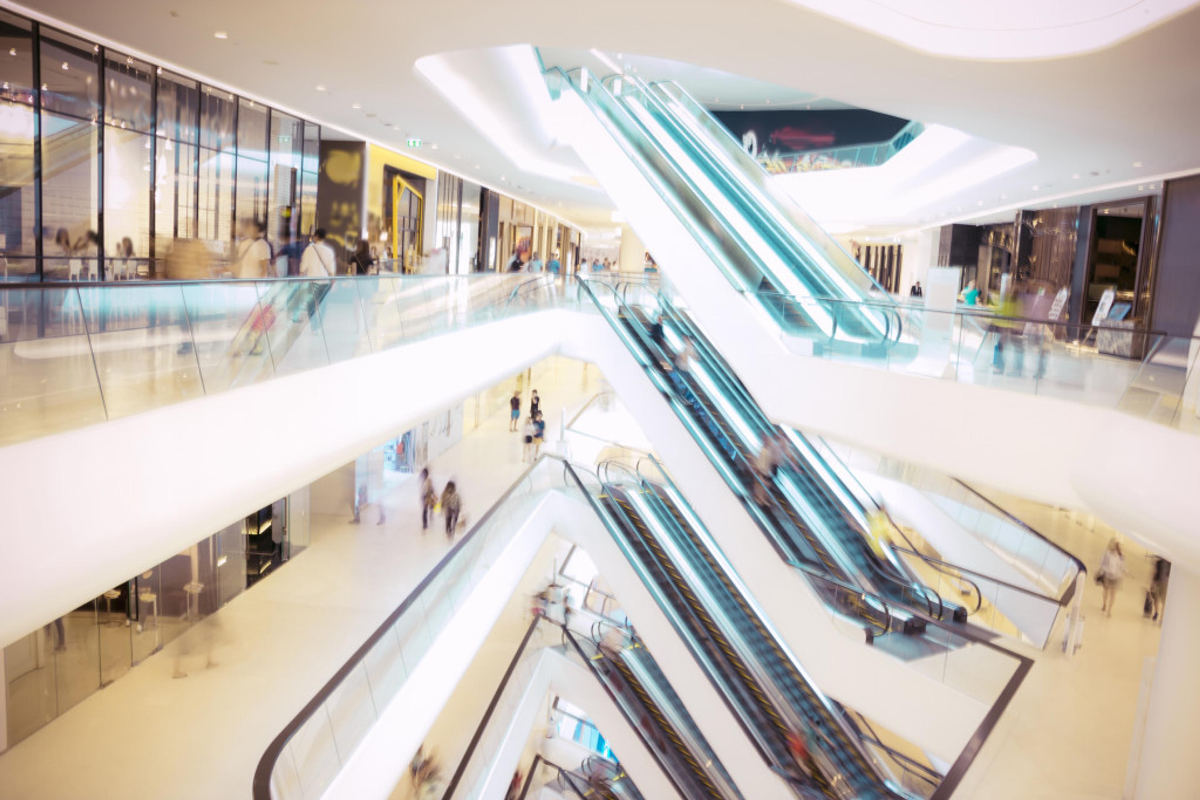 A modern shopping mall interior featuring multiple escalators and a bright, open design. Shoppers are seen moving throughout the space.