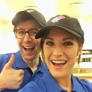 Two employees in blue uniforms and black caps smile at the camera, one giving a thumbs up. They appear to be in a restaurant setting.