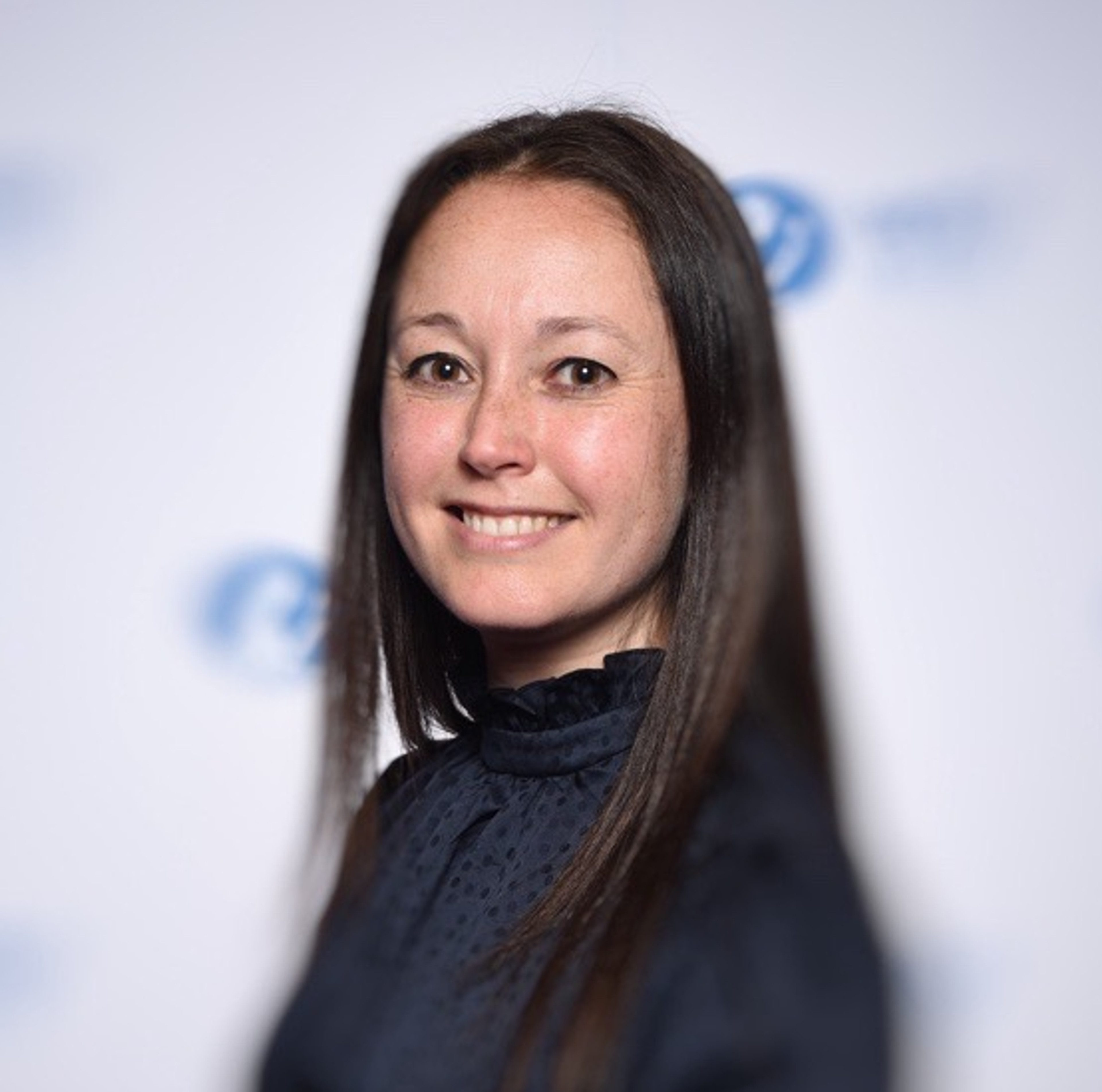 A woman with long brown hair smiles at the camera, wearing a dark blouse. The background features a blurred logo and soft lighting.