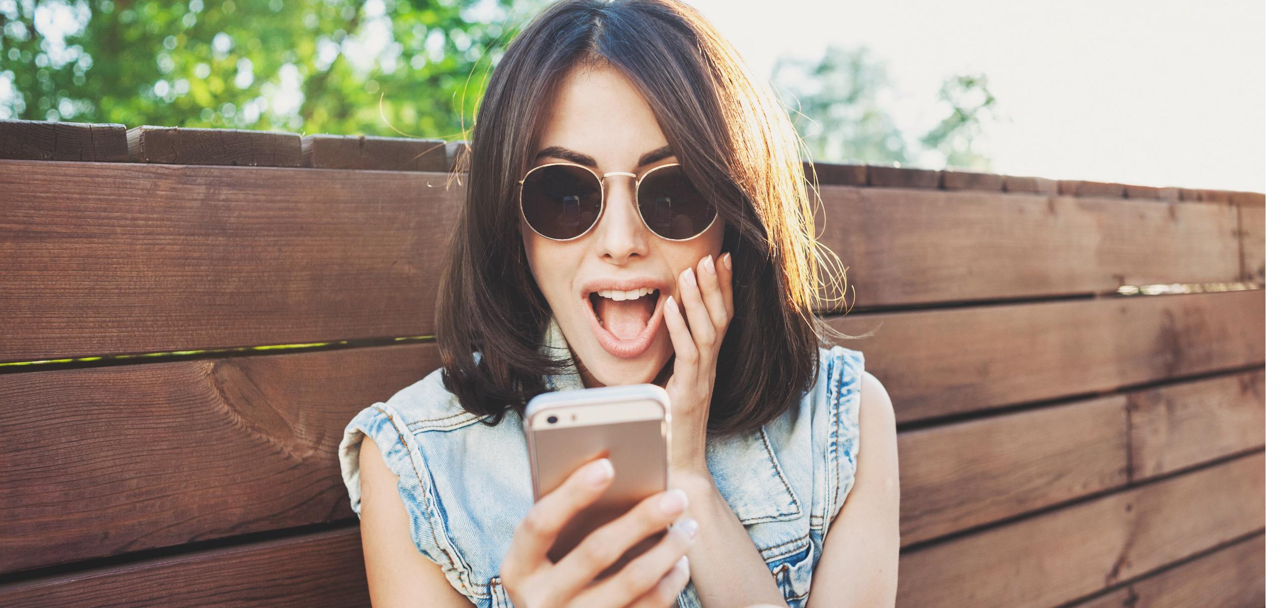 A young woman wearing sunglasses is sitting on a wooden bench, looking surprised while holding a smartphone. She has shoulder-length hair and is wearing a denim vest.