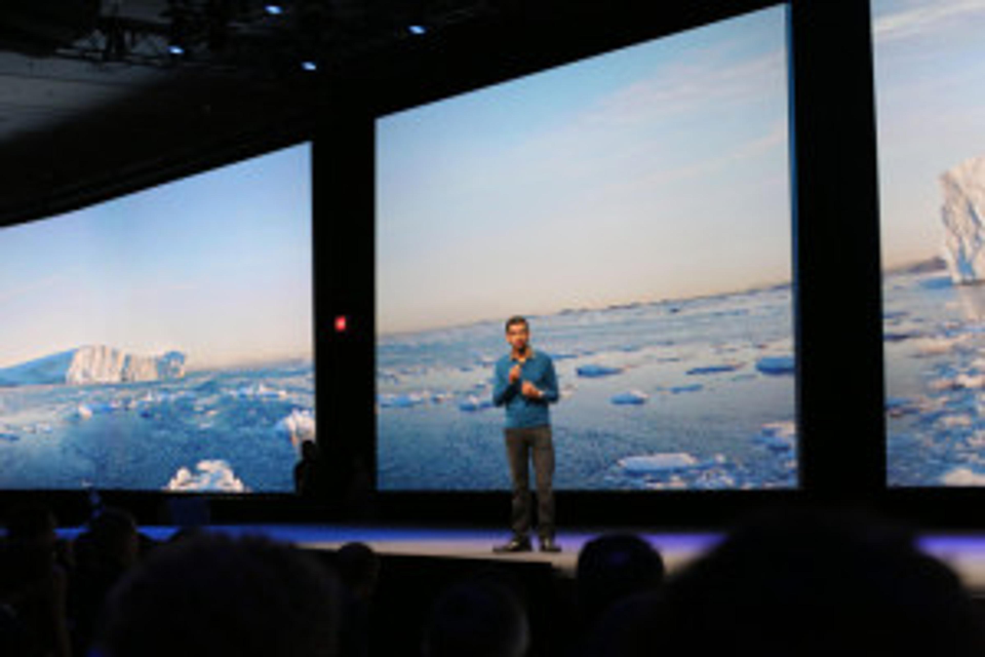 A speaker stands on stage in front of large screens displaying a serene icy landscape. The audience is visible in the foreground.