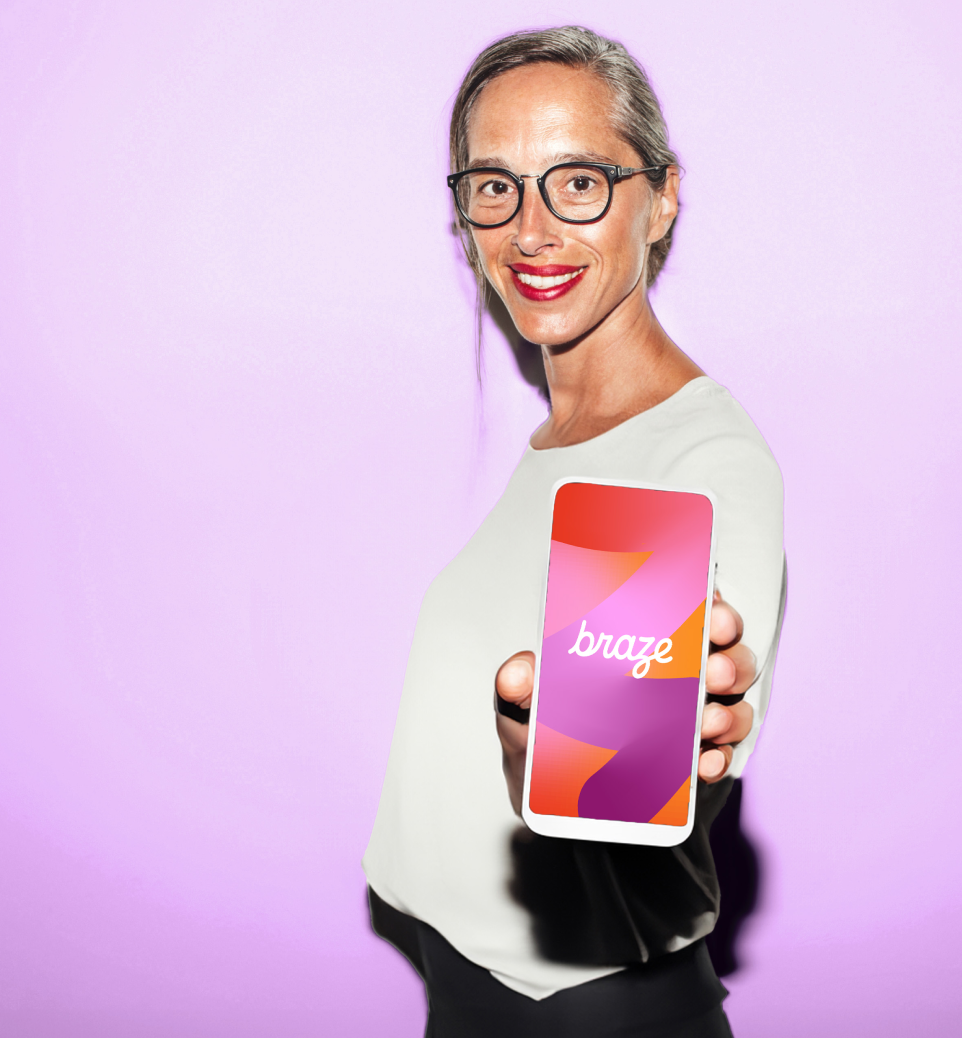 Person in glasses smiles and holds a smartphone toward the camera, displaying a colorful screen with the Braze logo against a light purple background.