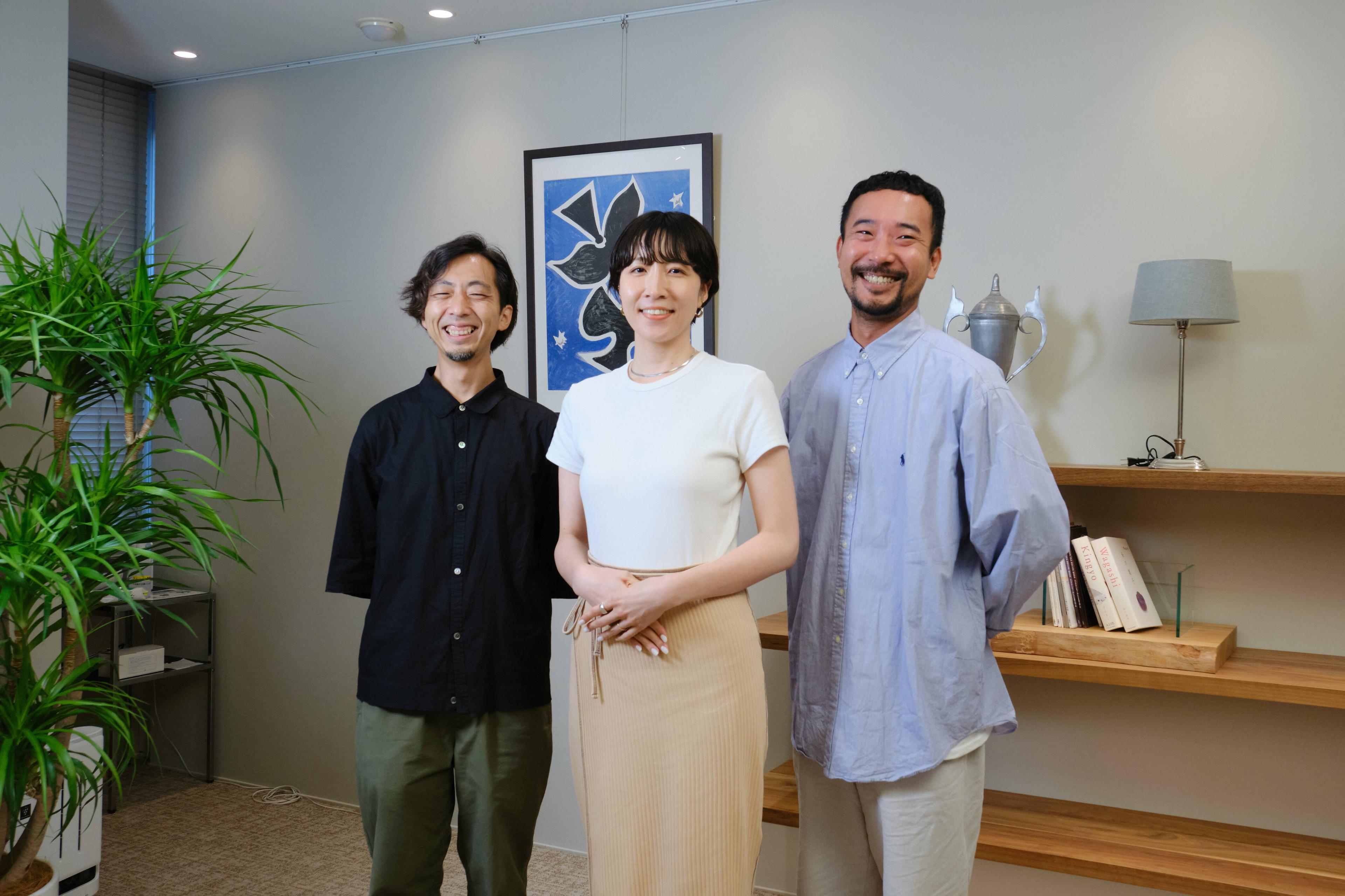 3人の人物が室内で笑顔で立っている。背景には植物とアート作品がある。