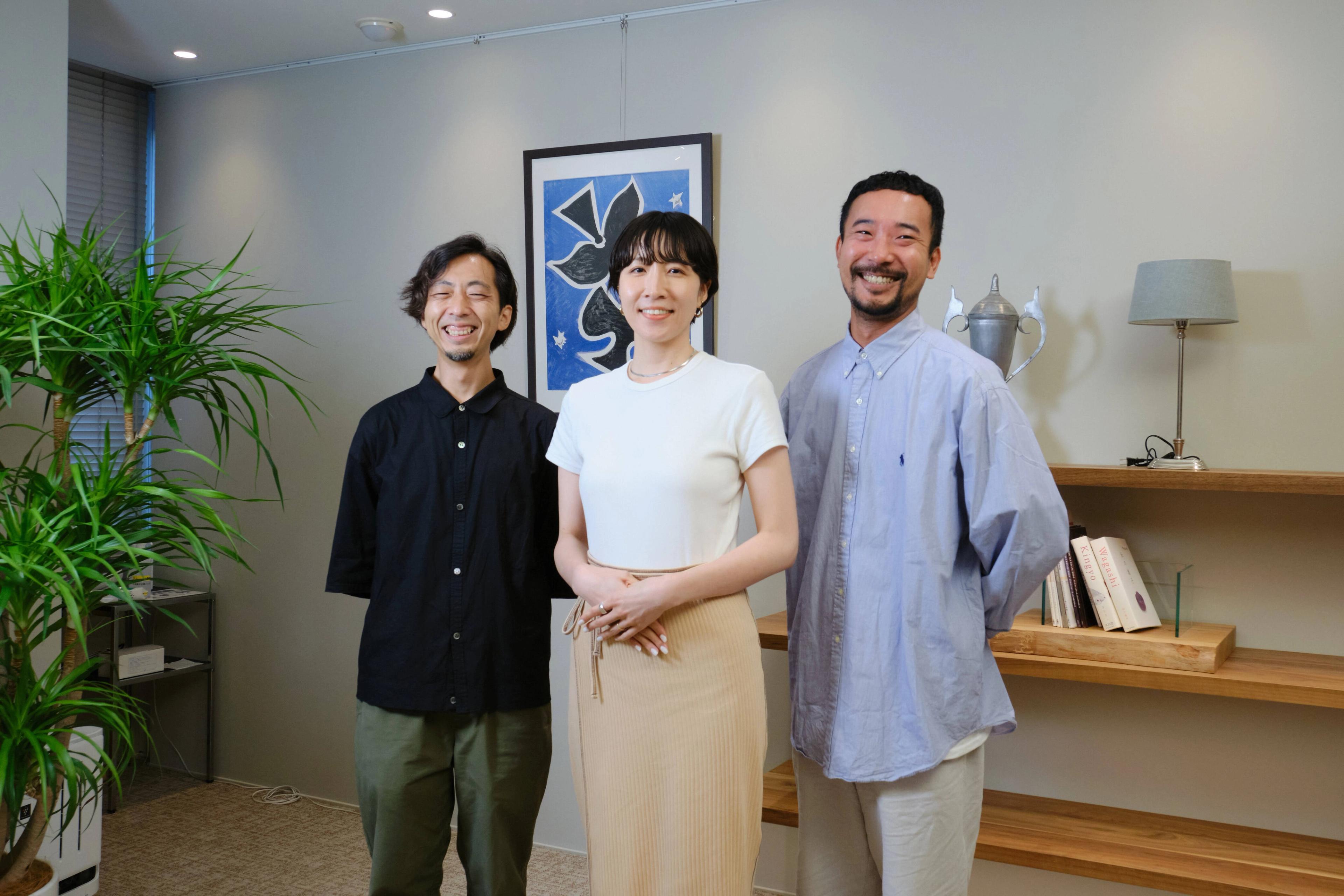 3人の人物が室内で笑顔で立っている。背景には植物とアート作品がある。