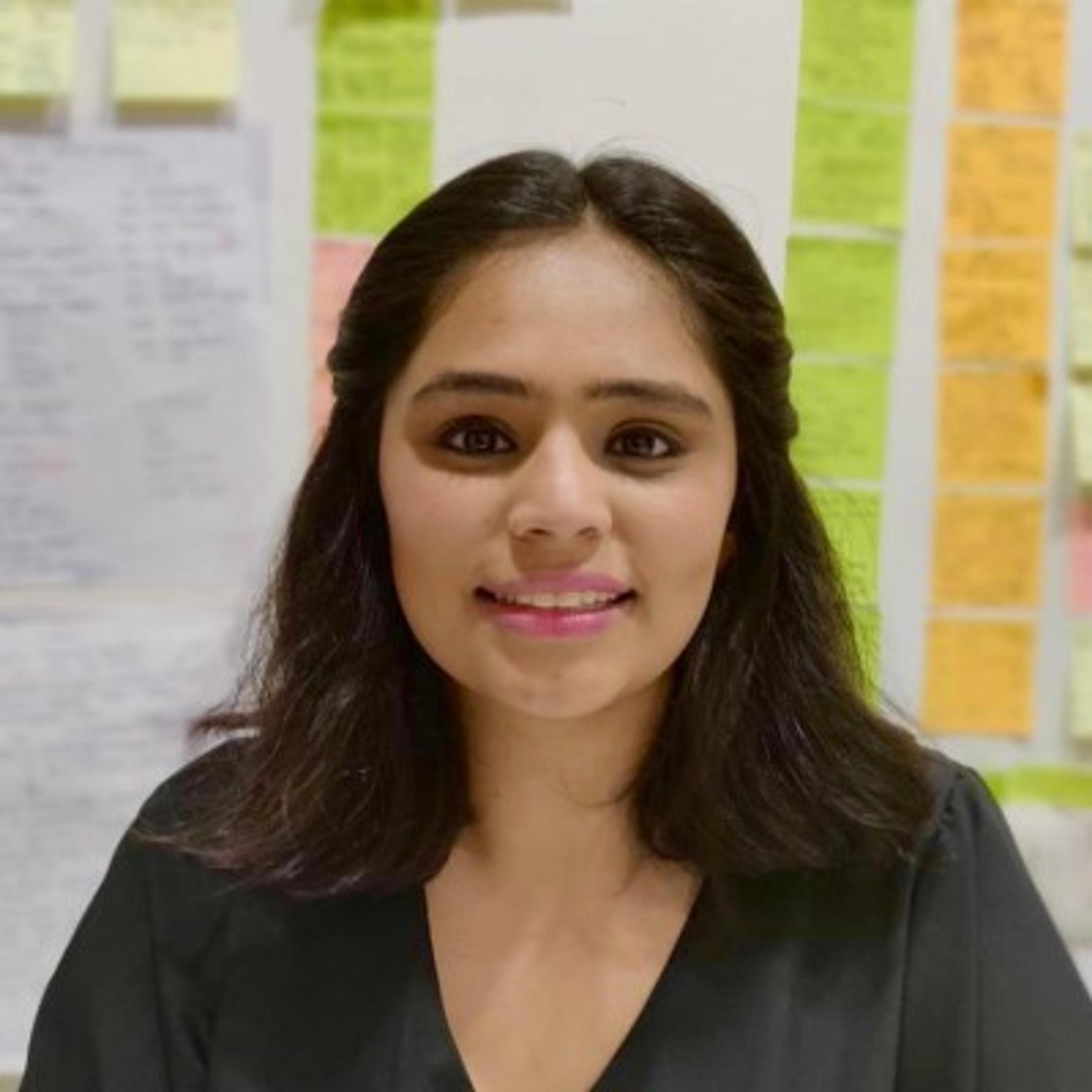 A woman with shoulder-length dark hair smiles at the camera, standing in front of a wall covered with colorful sticky notes. She is wearing a black top.