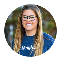 A smiling young woman with long hair and glasses is wearing a blue shirt that says "Neighbors." She is standing outdoors with a blurred background of greenery.