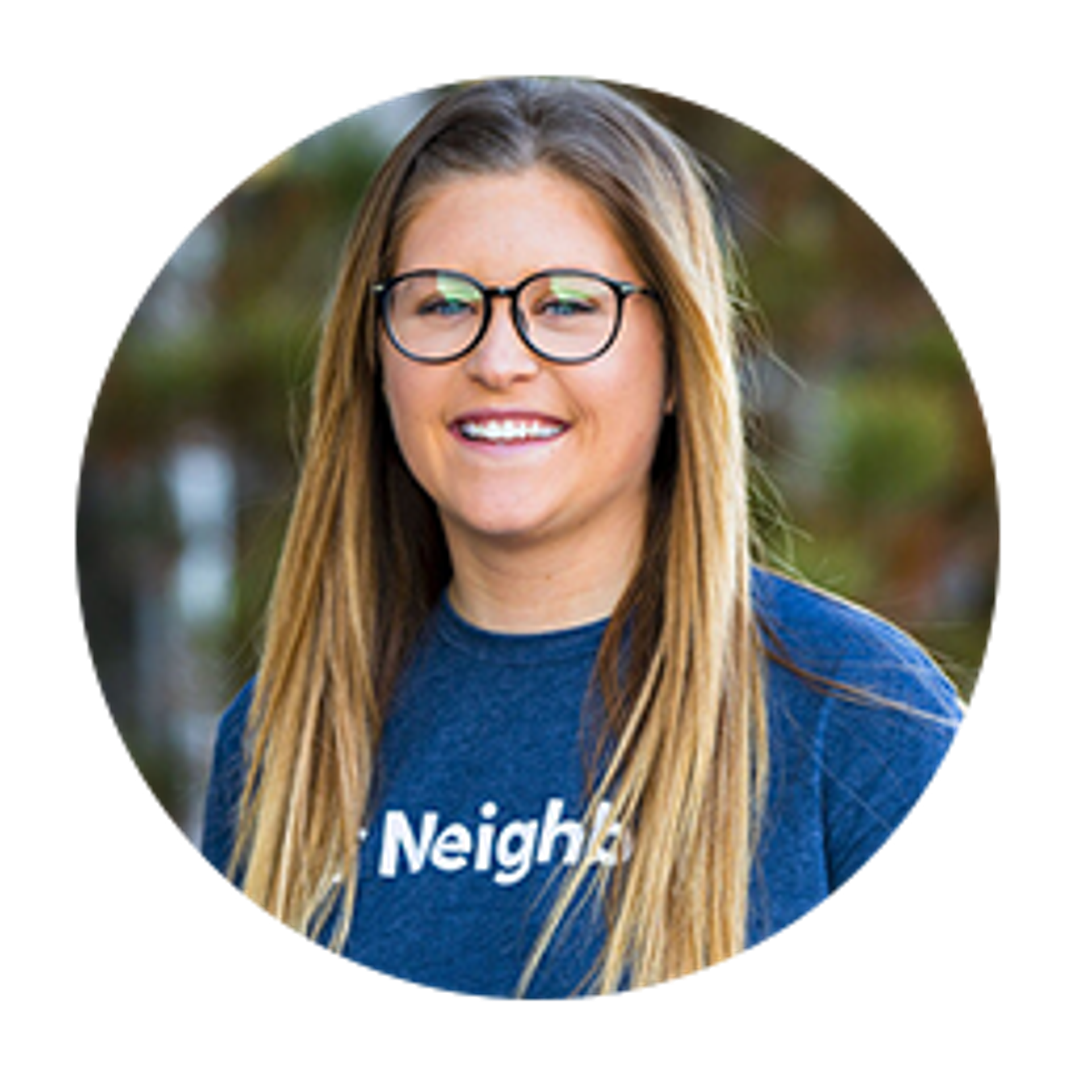 A smiling young woman with long hair and glasses is wearing a blue shirt that says "Neighbors." She is standing outdoors with a blurred background of greenery.
