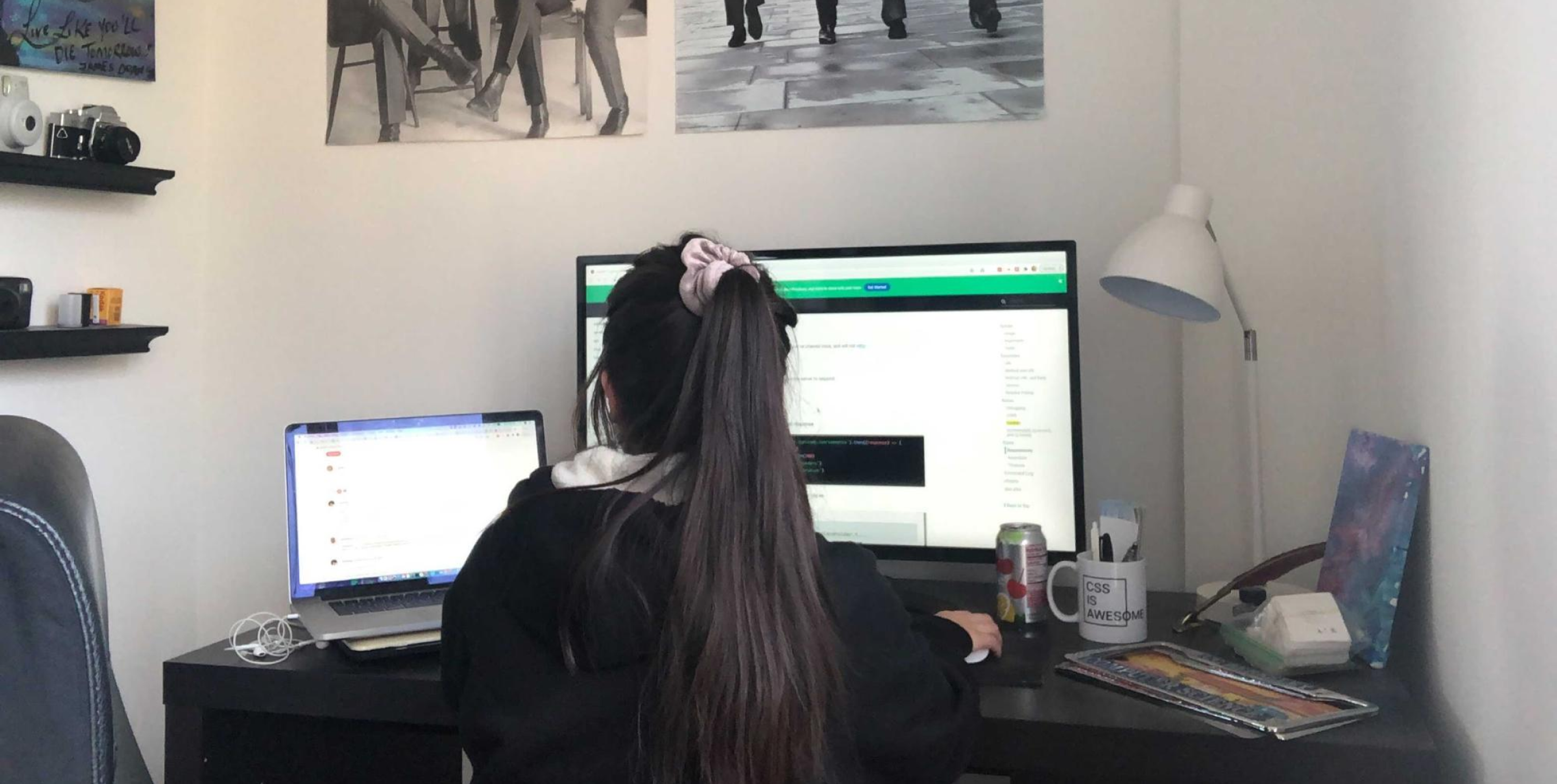 A person with long hair tied in a ponytail sits at a desk, working on a computer with a second laptop nearby. The wall features black-and-white photographs.
