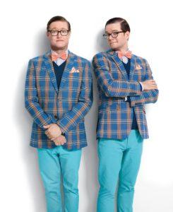 Two men in matching outfits featuring plaid blazers and bright blue pants, standing side by side with different poses. Both wear glasses and bow ties.