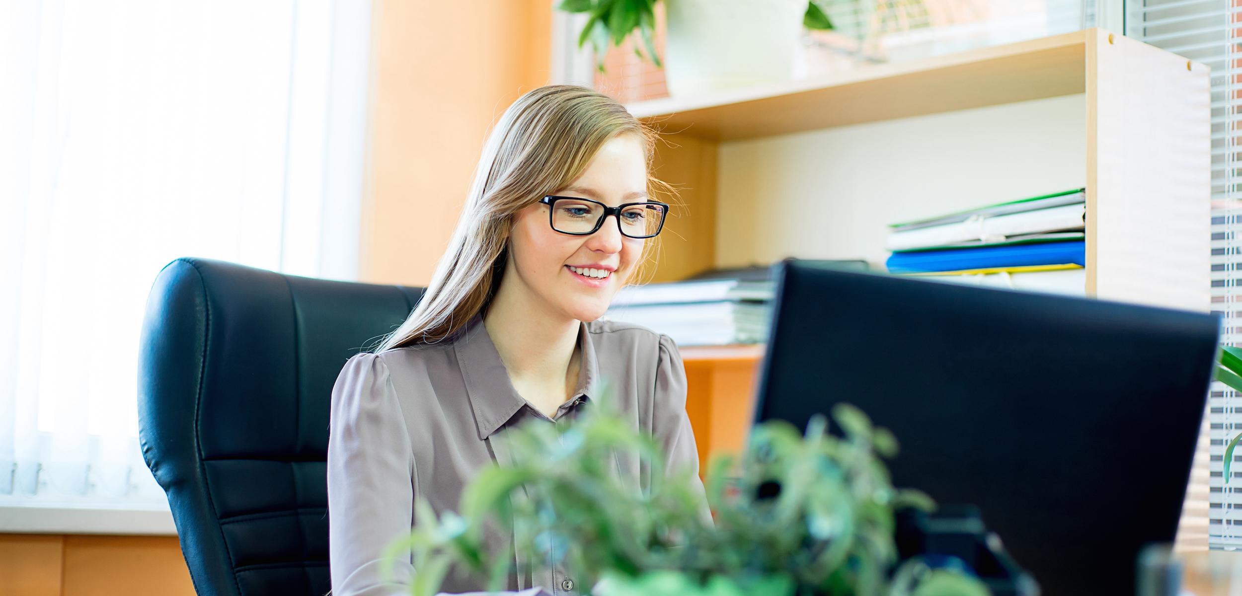 A woman with glasses smiles while working at a desk with a computer. Green plants are visible in the foreground and background.