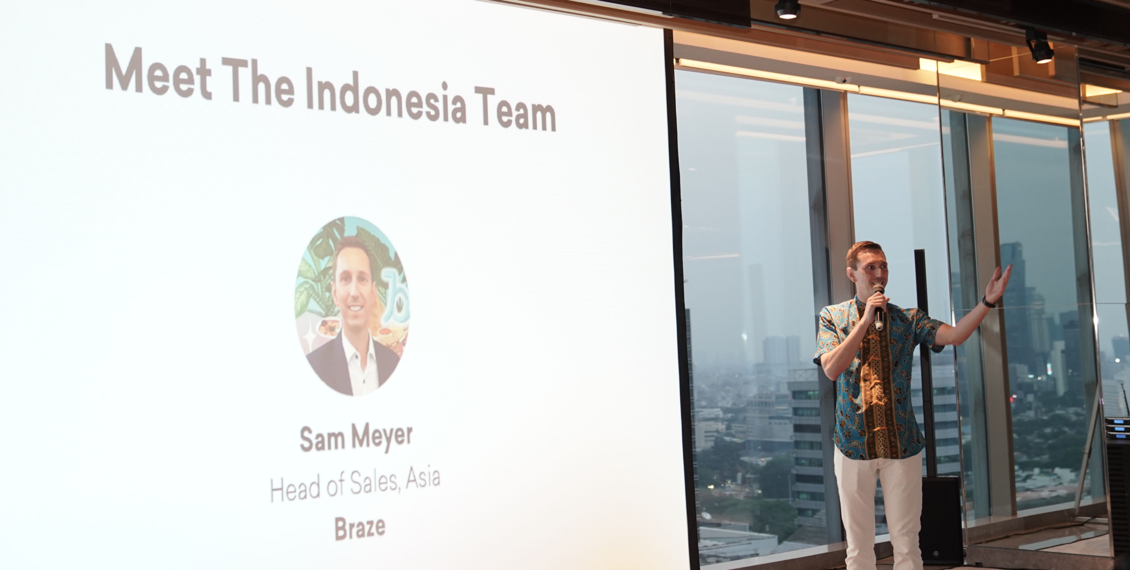 A man in a colorful shirt speaks to an audience in front of a presentation slide introducing Sam Meyer, Head of Sales, Asia at Braze. The city skyline is visible through large windows behind him.