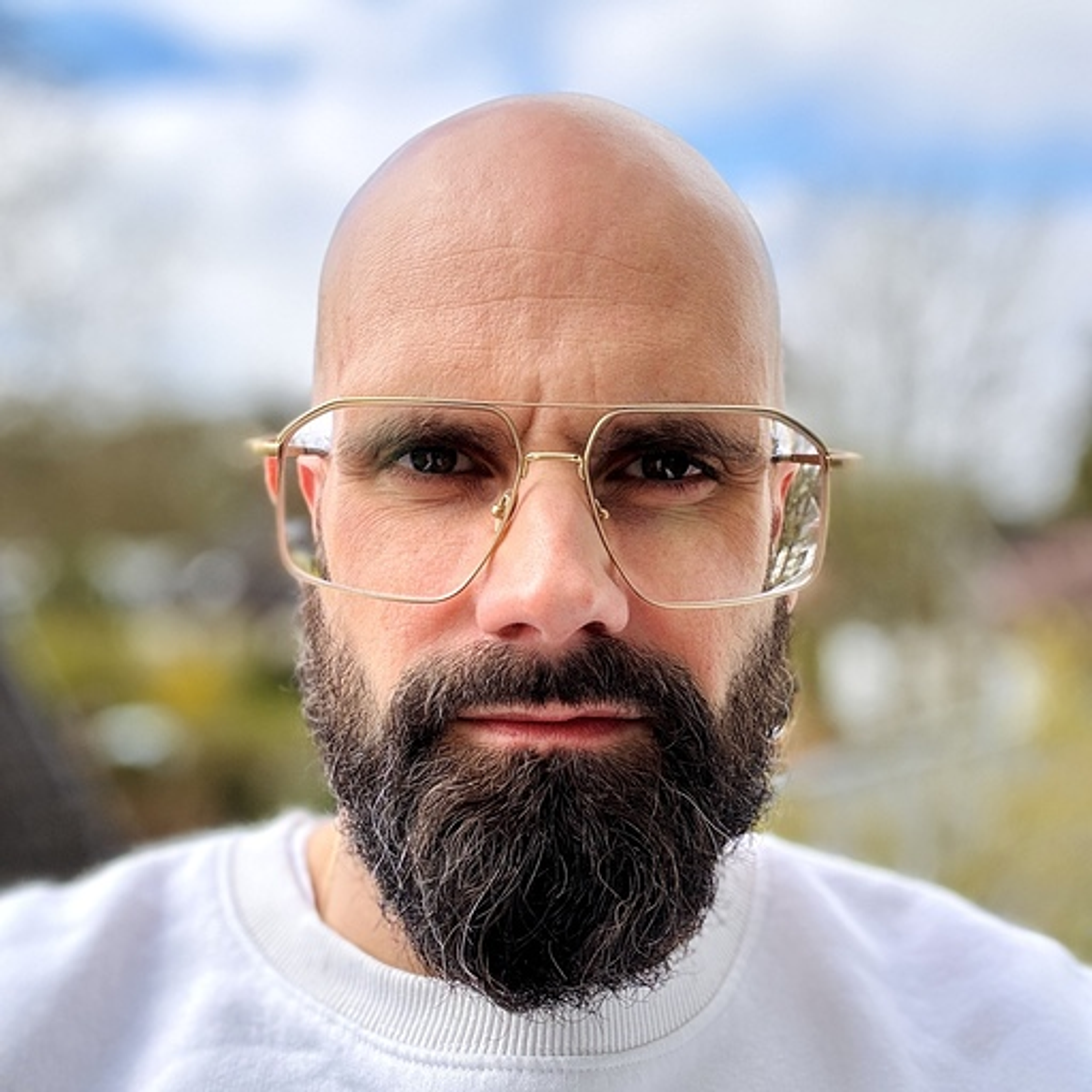 Bald man with with a full dark beard and large clear-framed glasses looking directly at the camera outdoors