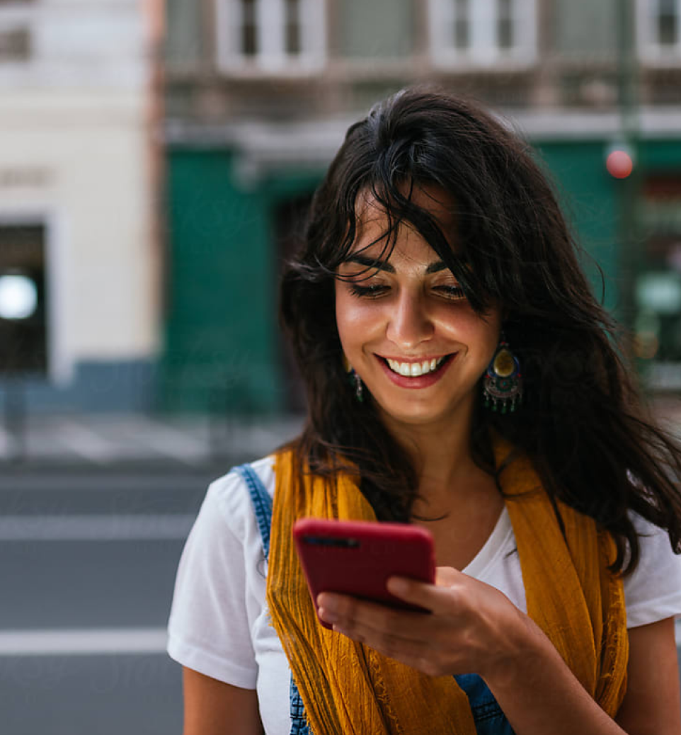 Mulher sorridente com cabelo solto, usando uma blusa branca e um lenço amarelo, olhando para o celular. Ao fundo, uma rua urbana desfocada.