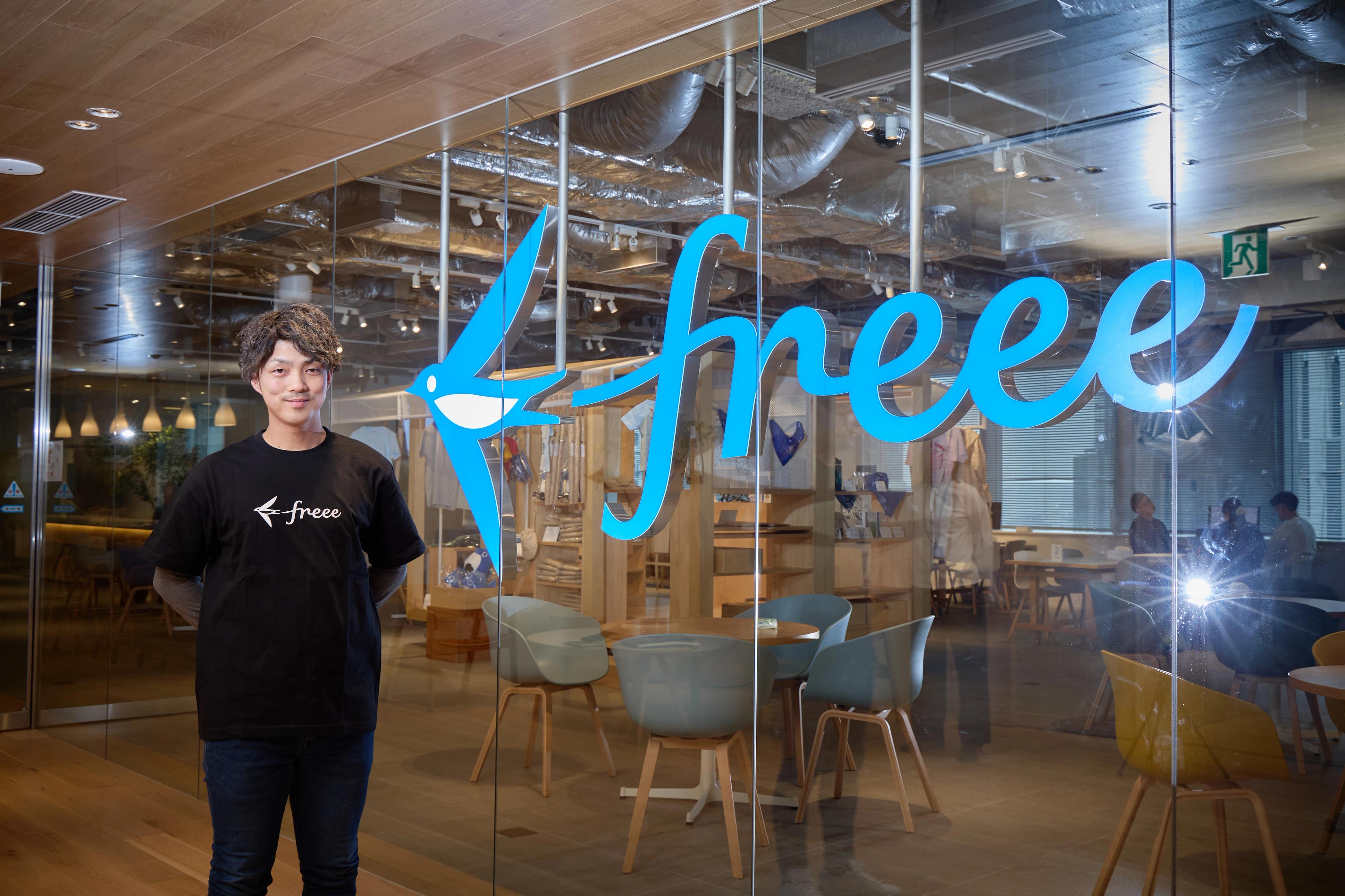 黒い「freee」のTシャツを着た男性が、大きな青い「freee」のロゴが入ったガラス壁の前で微笑み、その向こうにはオフィスの内部が見えている。