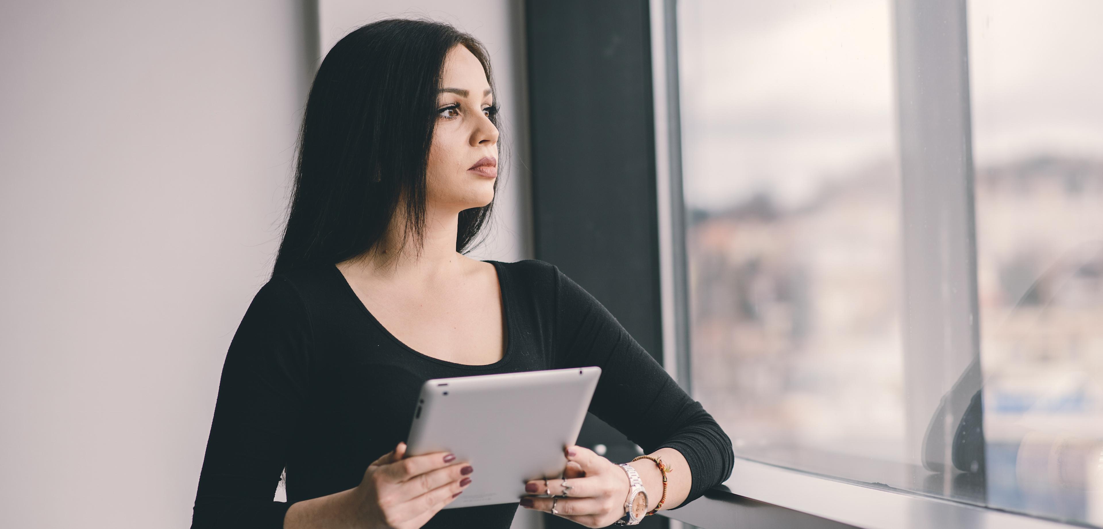A woman with long dark hair stands by a window, holding a tablet and looking thoughtfully outside. She is wearing a black top and appears contemplative.
