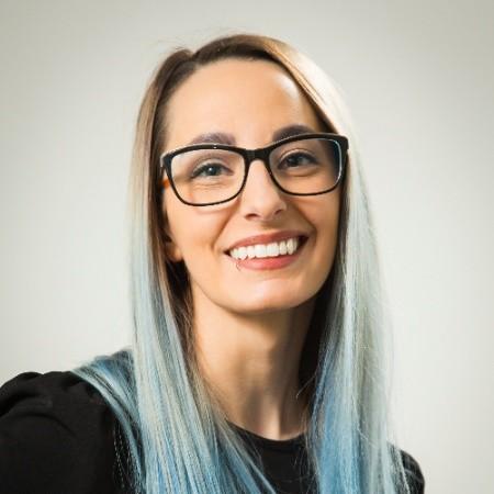 A woman with long, light blue hair and glasses smiles at the camera. She is wearing a black top against a light background.
