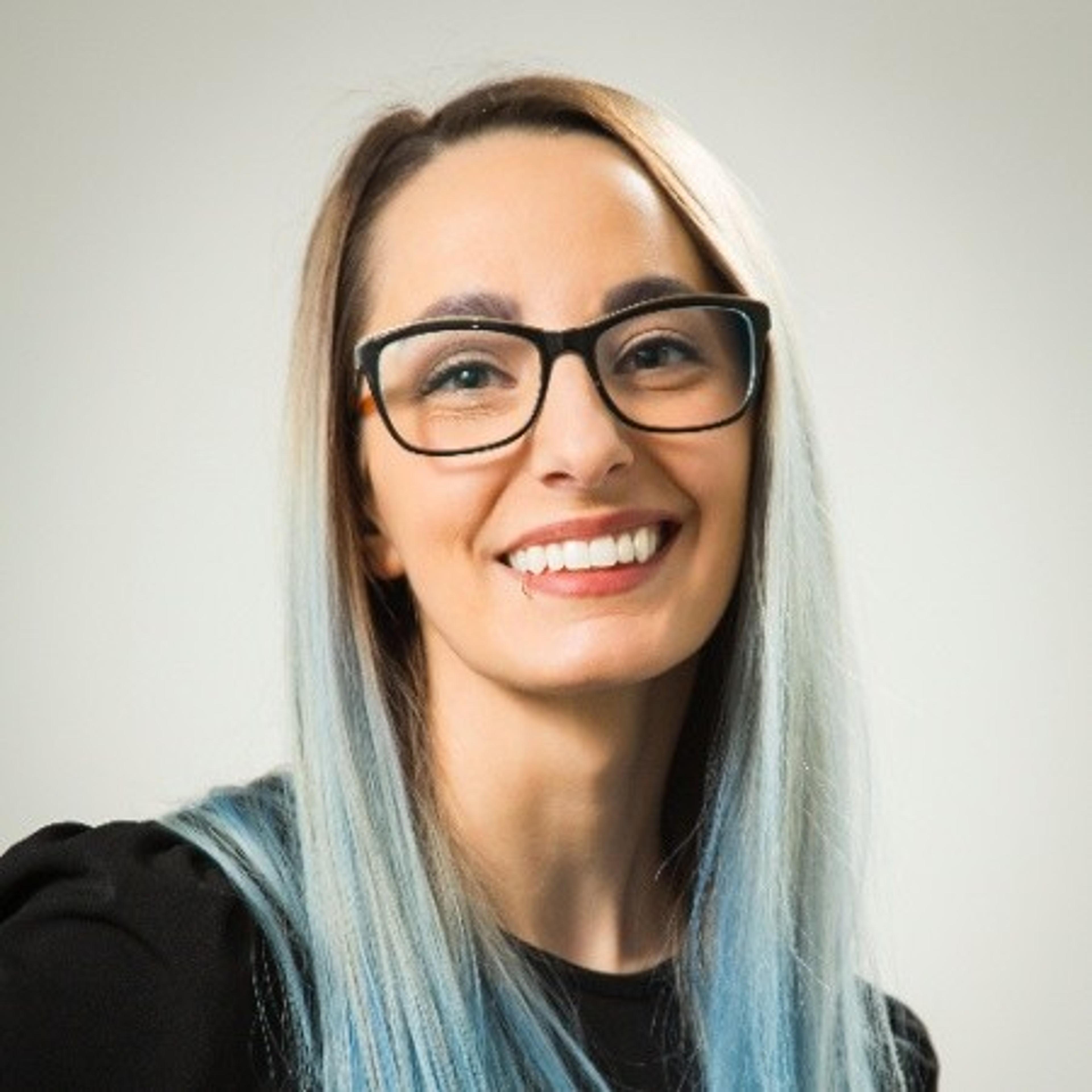 A woman with long, light blue hair and glasses smiles at the camera. She is wearing a black top against a light background.