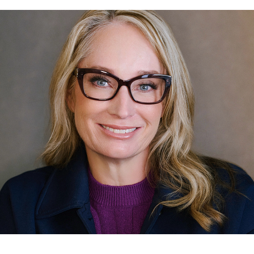 Headshot of a smiling blonde woman wearing dark-framed glasses, a purple top, and a navy blazer.