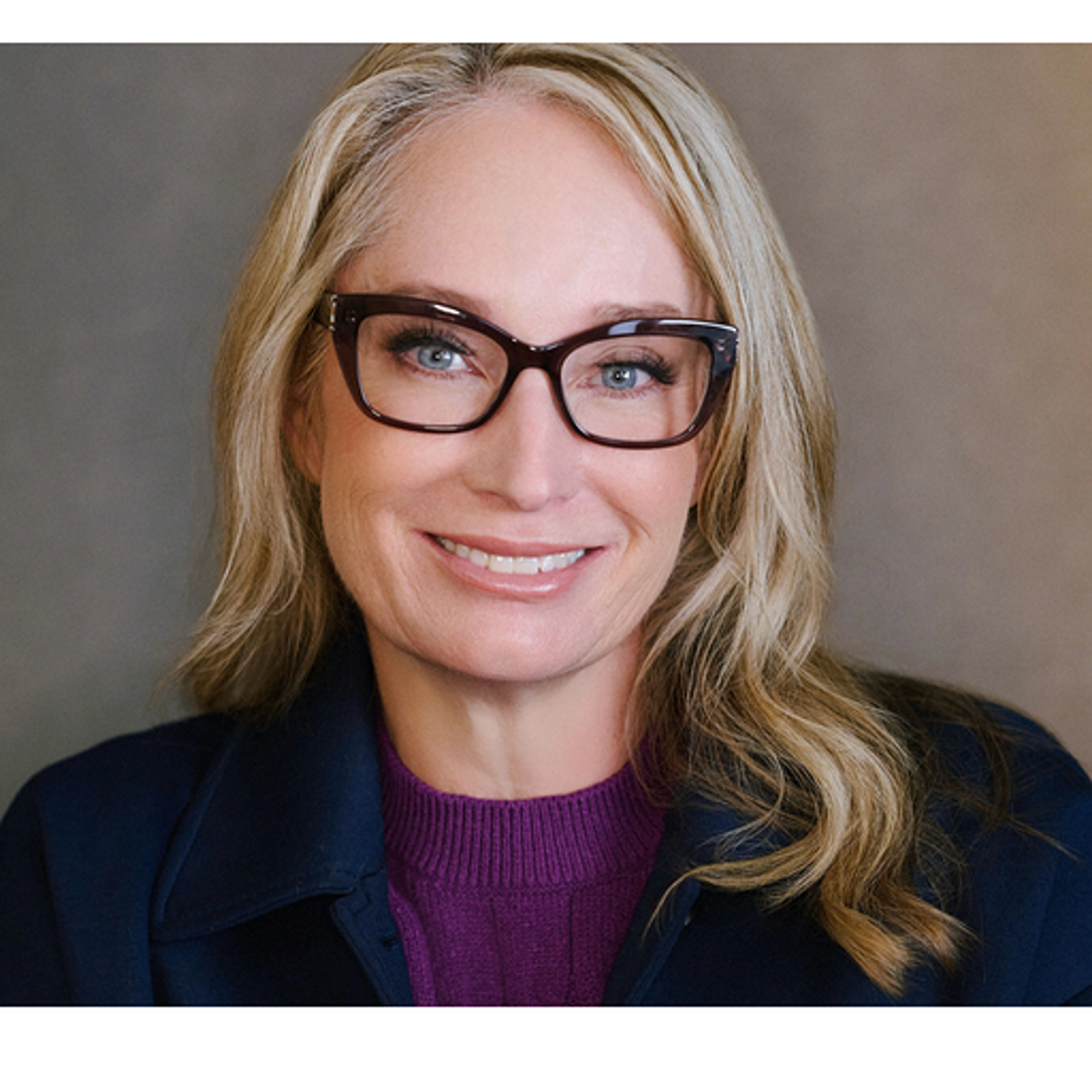 Headshot of a smiling blonde woman wearing dark-framed glasses, a purple top, and a navy blazer.