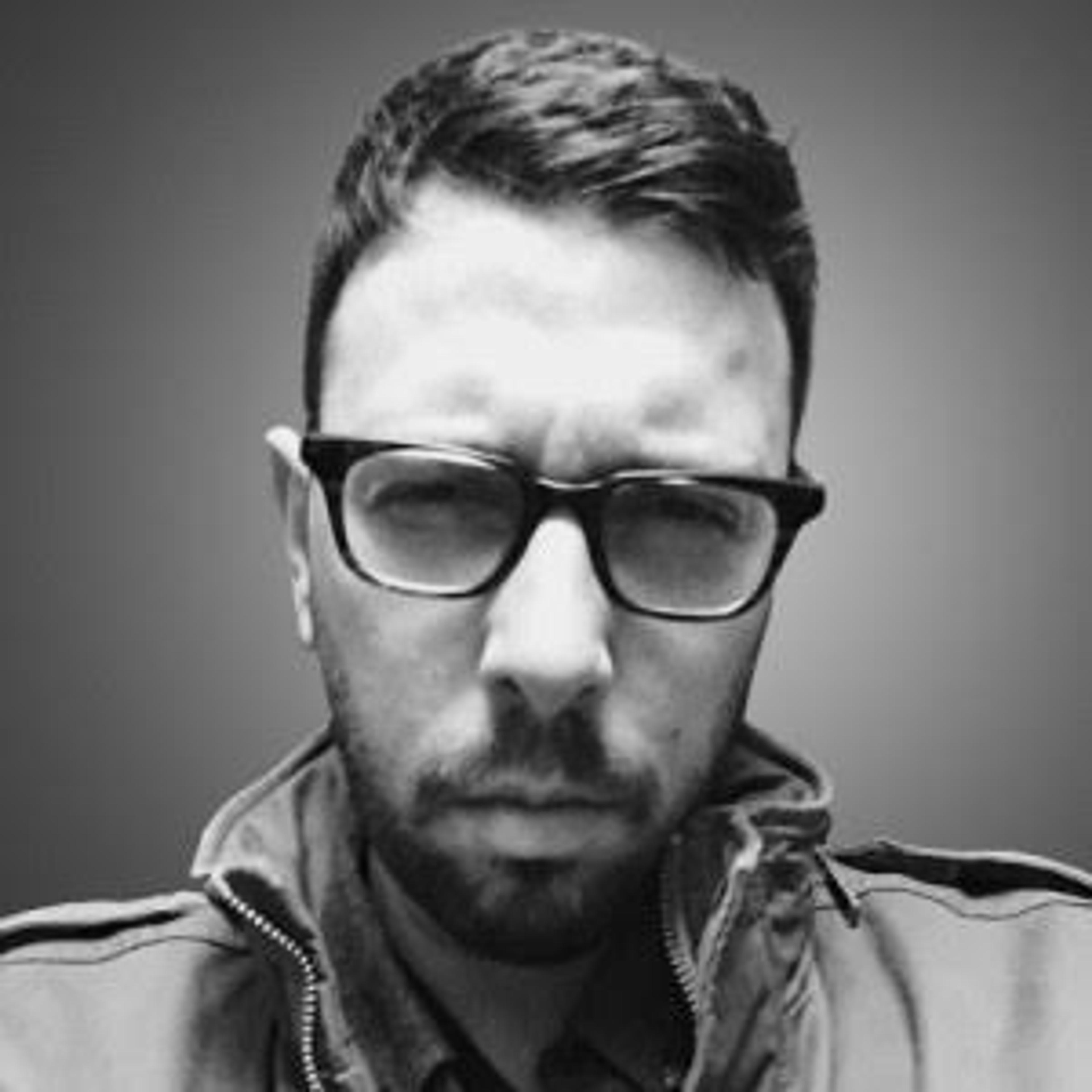 A man with glasses and a serious expression is looking directly at the camera. He is wearing a jacket and has short hair.
