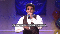 A man in a tuxedo performs on stage, singing about celebrating Hanukkah and dating Rihanna. Colorful balloons and a festive banner are in the background.