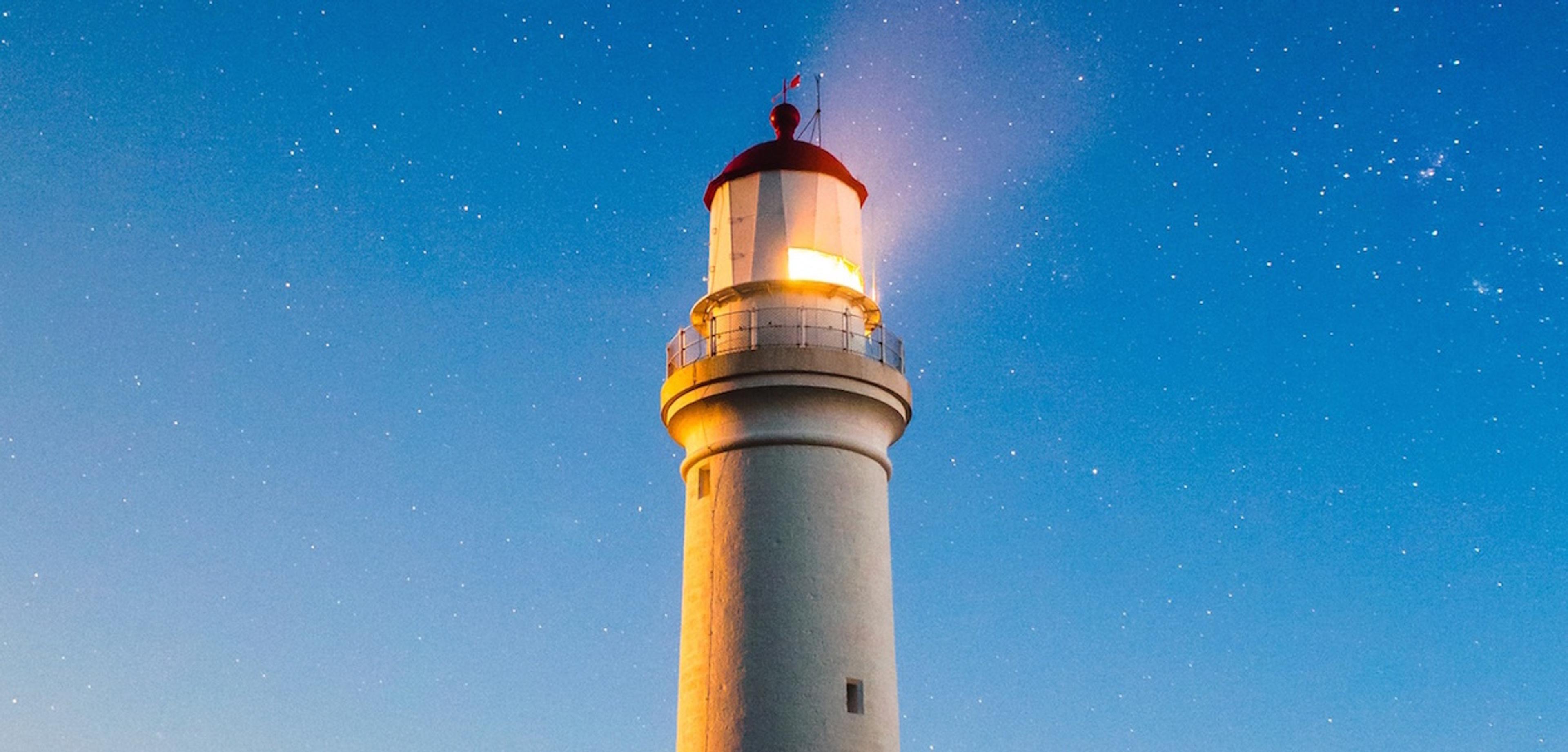 A tall lighthouse stands against a twilight sky, its light shining brightly. Stars are visible in the background.