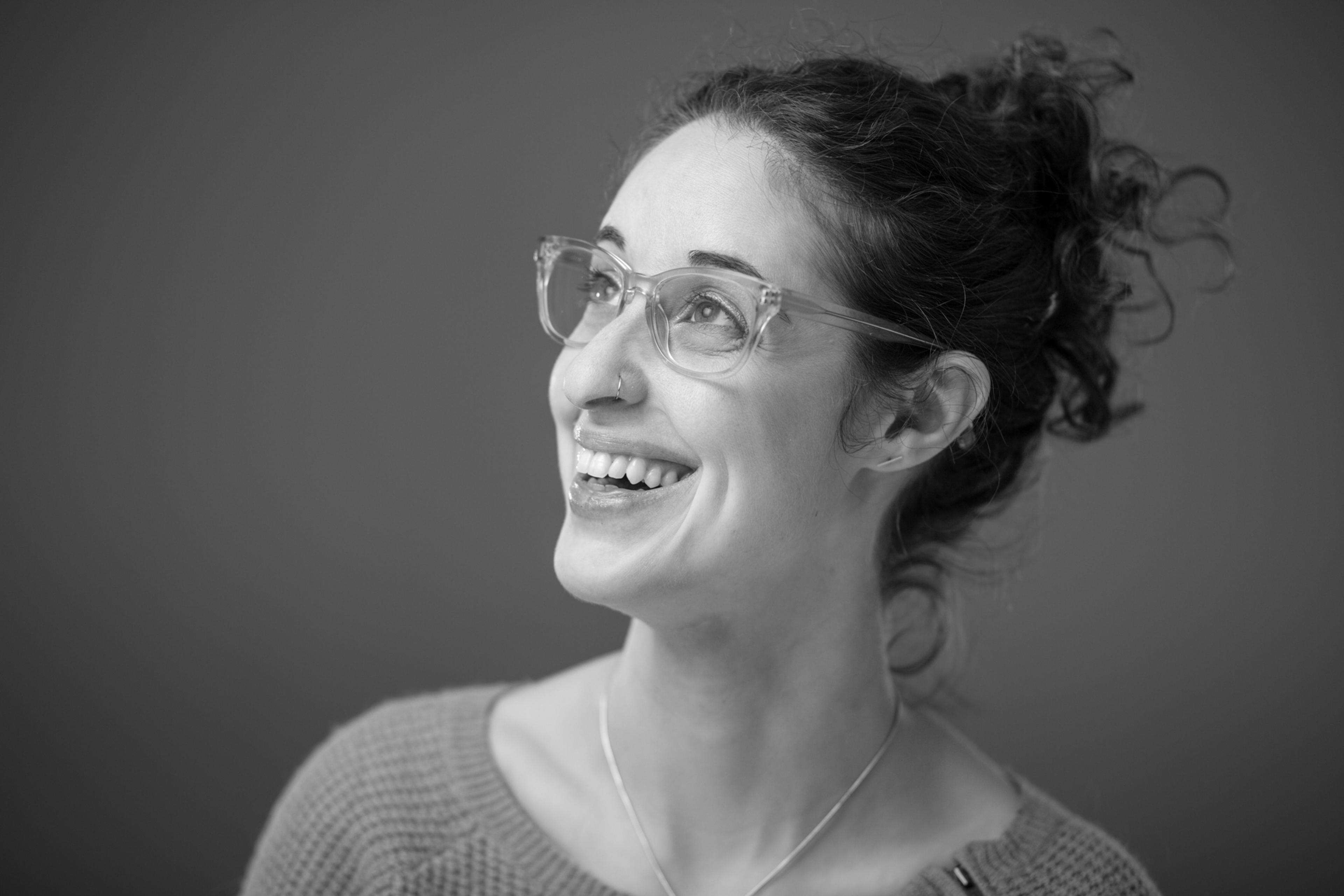A smiling woman with curly hair and glasses looks upward, wearing a cozy sweater. The image is in black and white.
