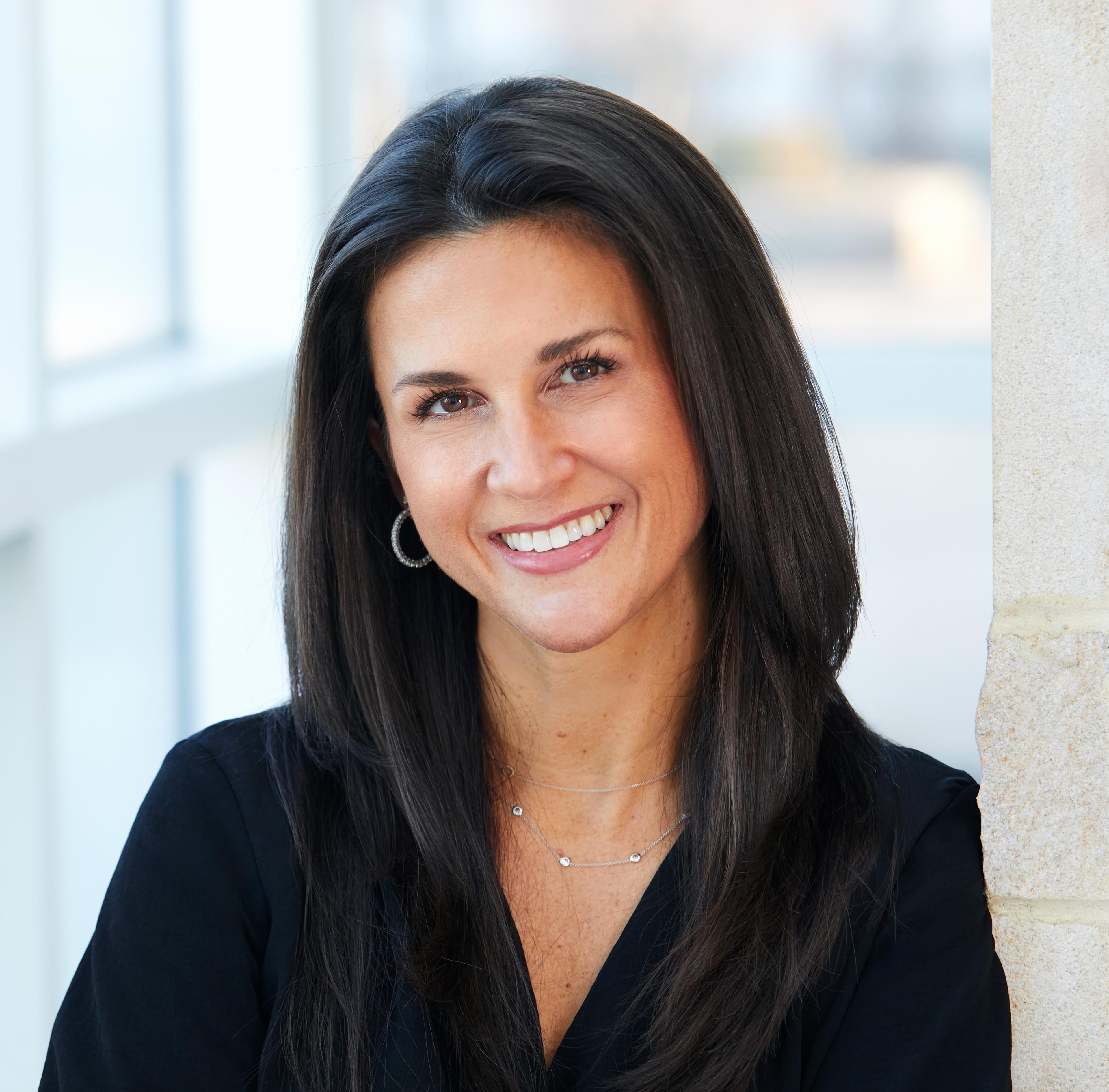 A woman with long dark hair smiles while leaning against a wall. She is wearing a black top and has hoop earrings.