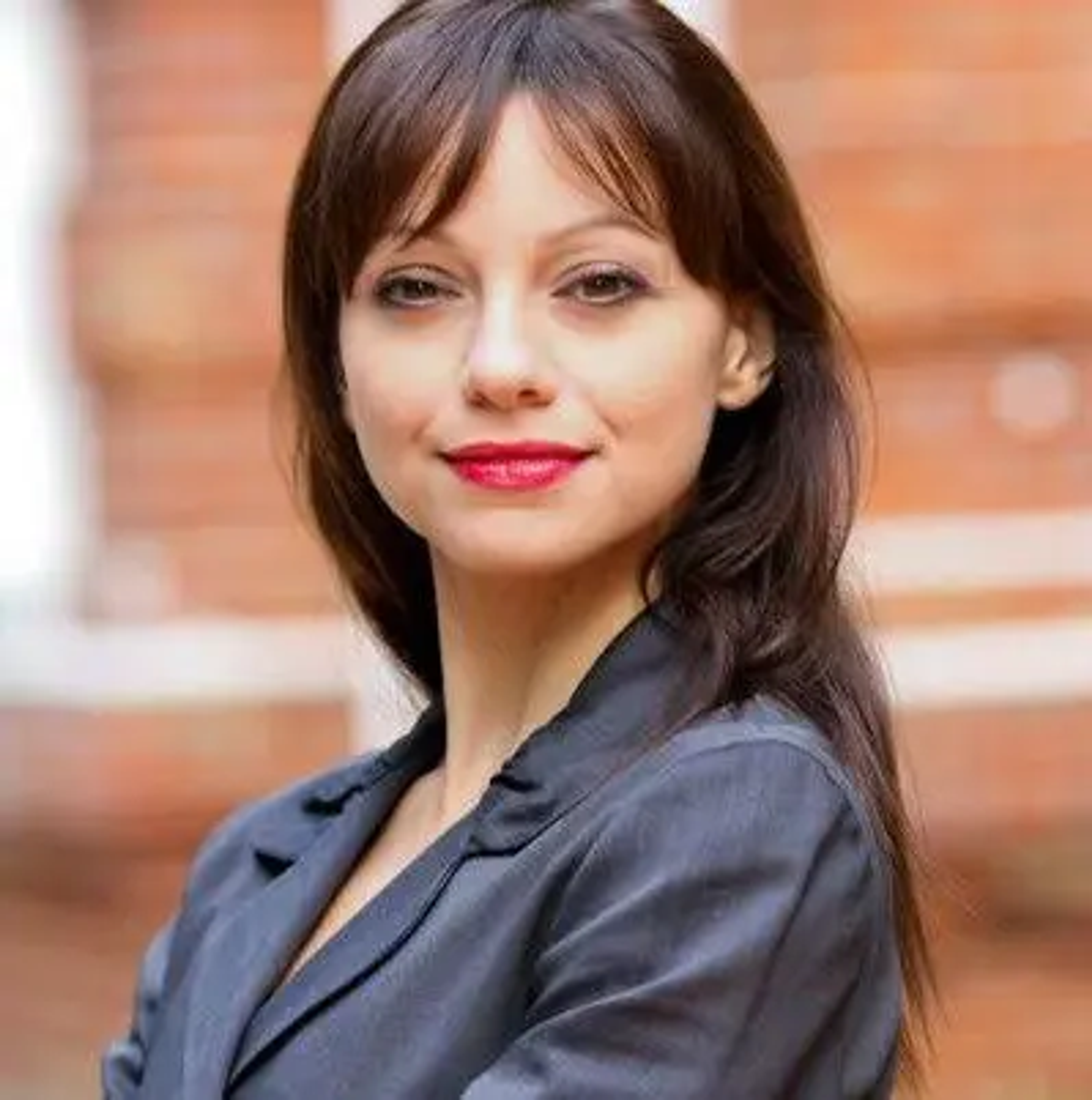 A woman with long dark hair and red lipstick poses confidently in a gray blazer against a blurred brick background. She has a slight smile and her arms are crossed.