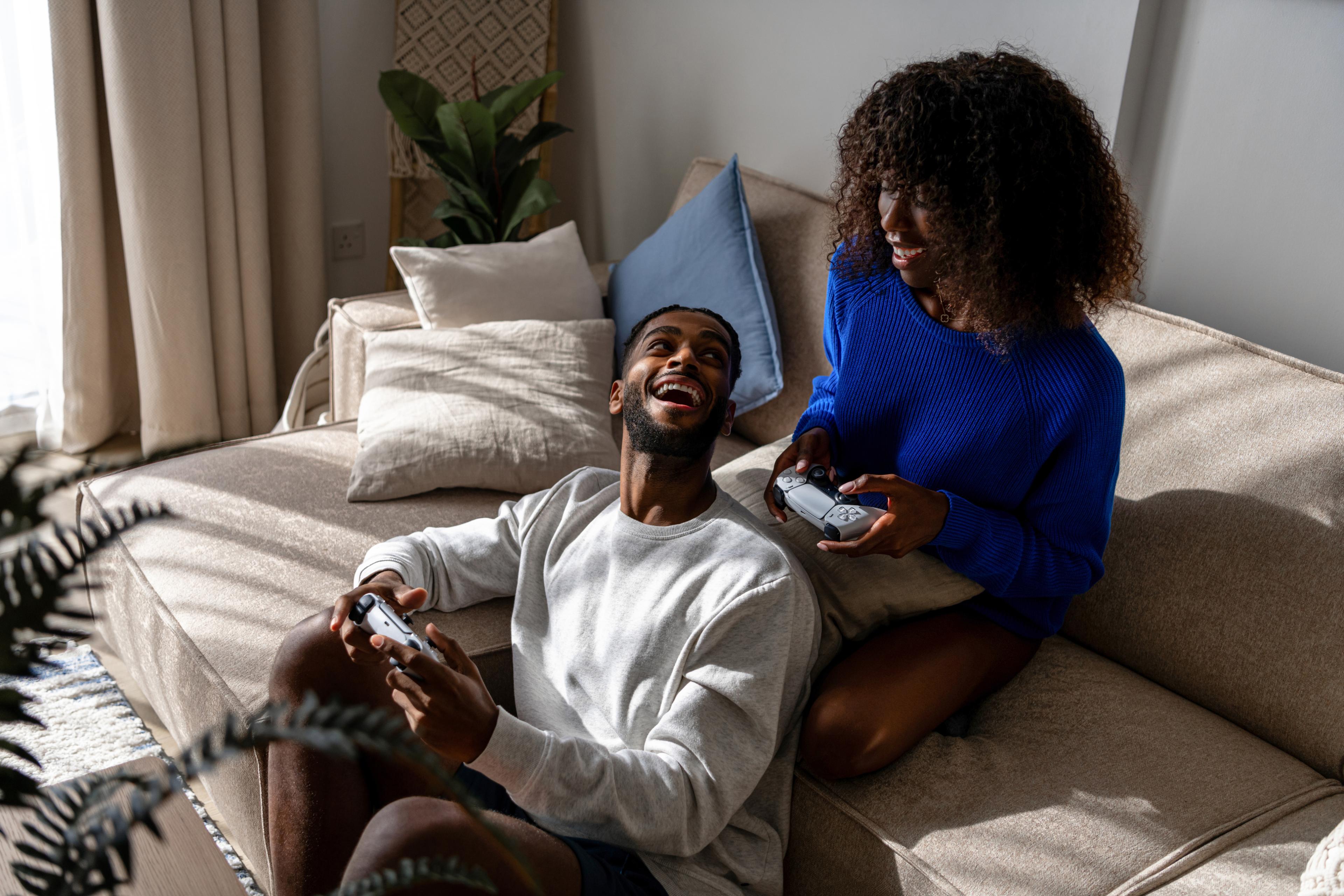 A man and woman are sitting on a couch, both holding video game controllers and smiling at each other. The room is bright with plants and cushions in the background.