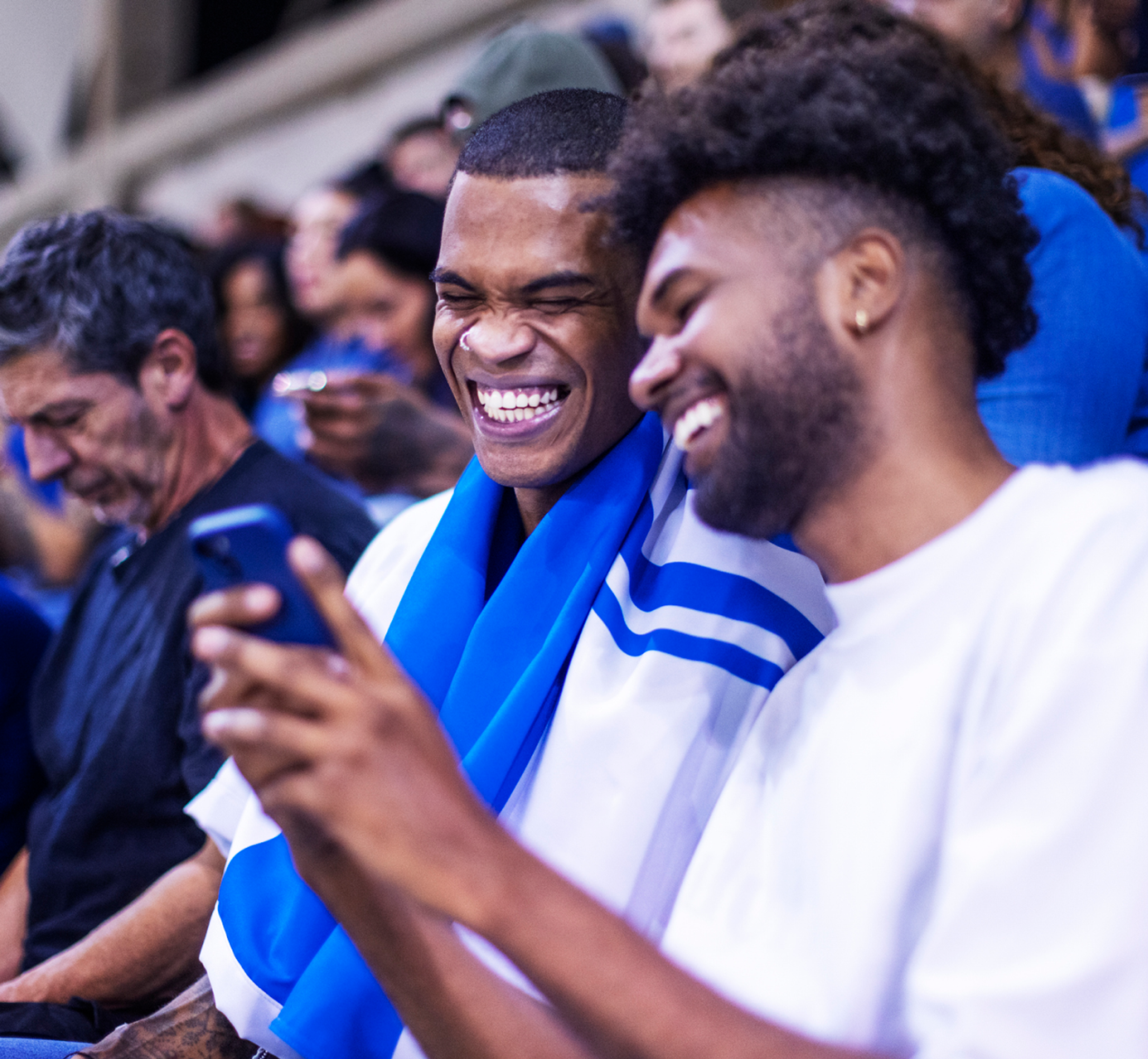 Two friends in a crowded stadium laugh together while looking at something on a smartphone, one draped in a blue and white fan scarf.
