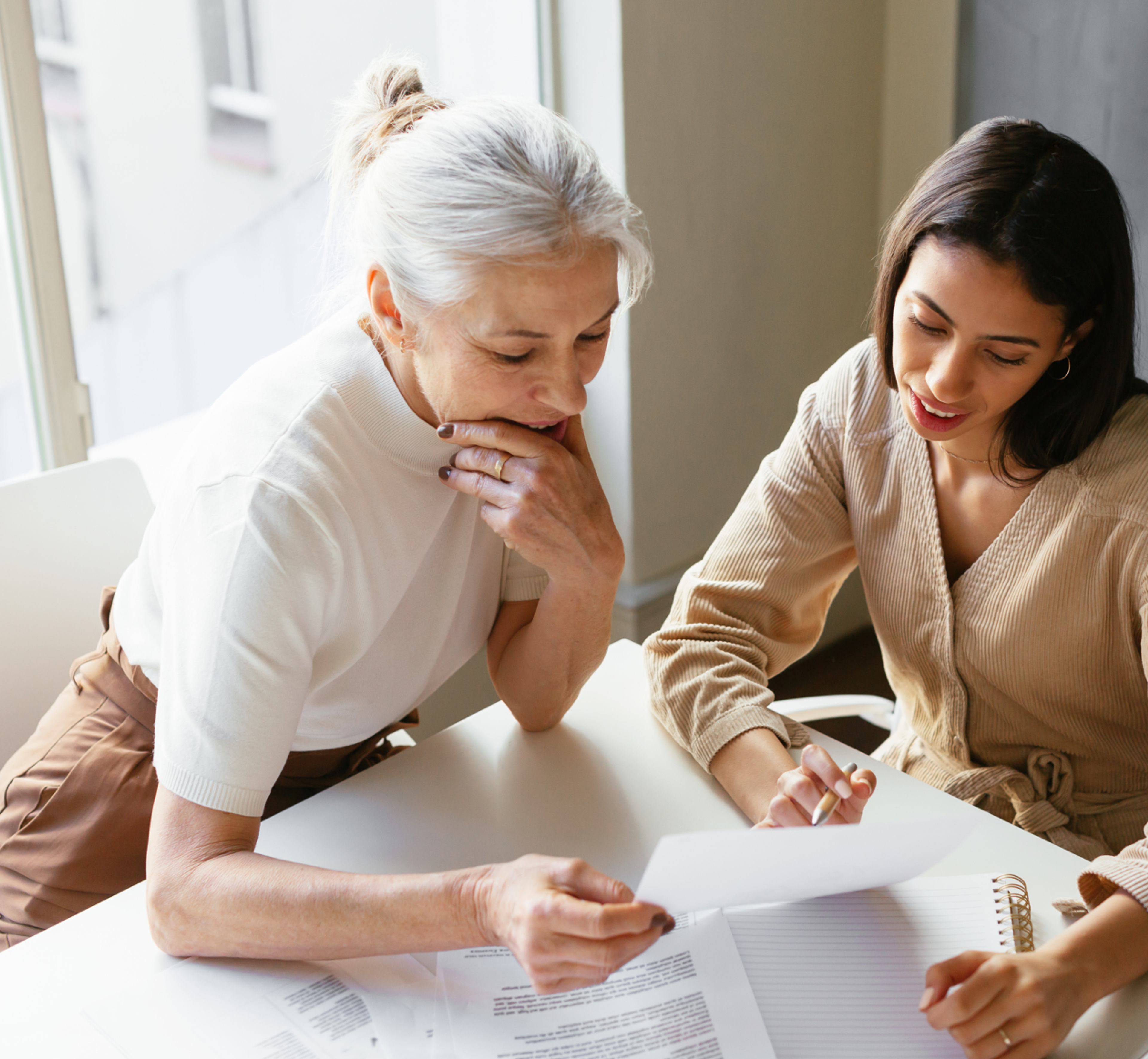 Two women sit at a table reviewing documents together, one holding a paper while the other takes notes in a notebook.