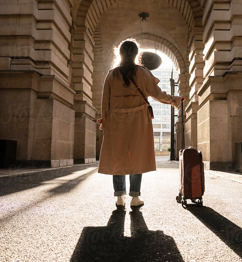 A person in a long coat stands under an archway, holding a hat and a suitcase. The sunlight creates a silhouette effect as they look out onto the street.