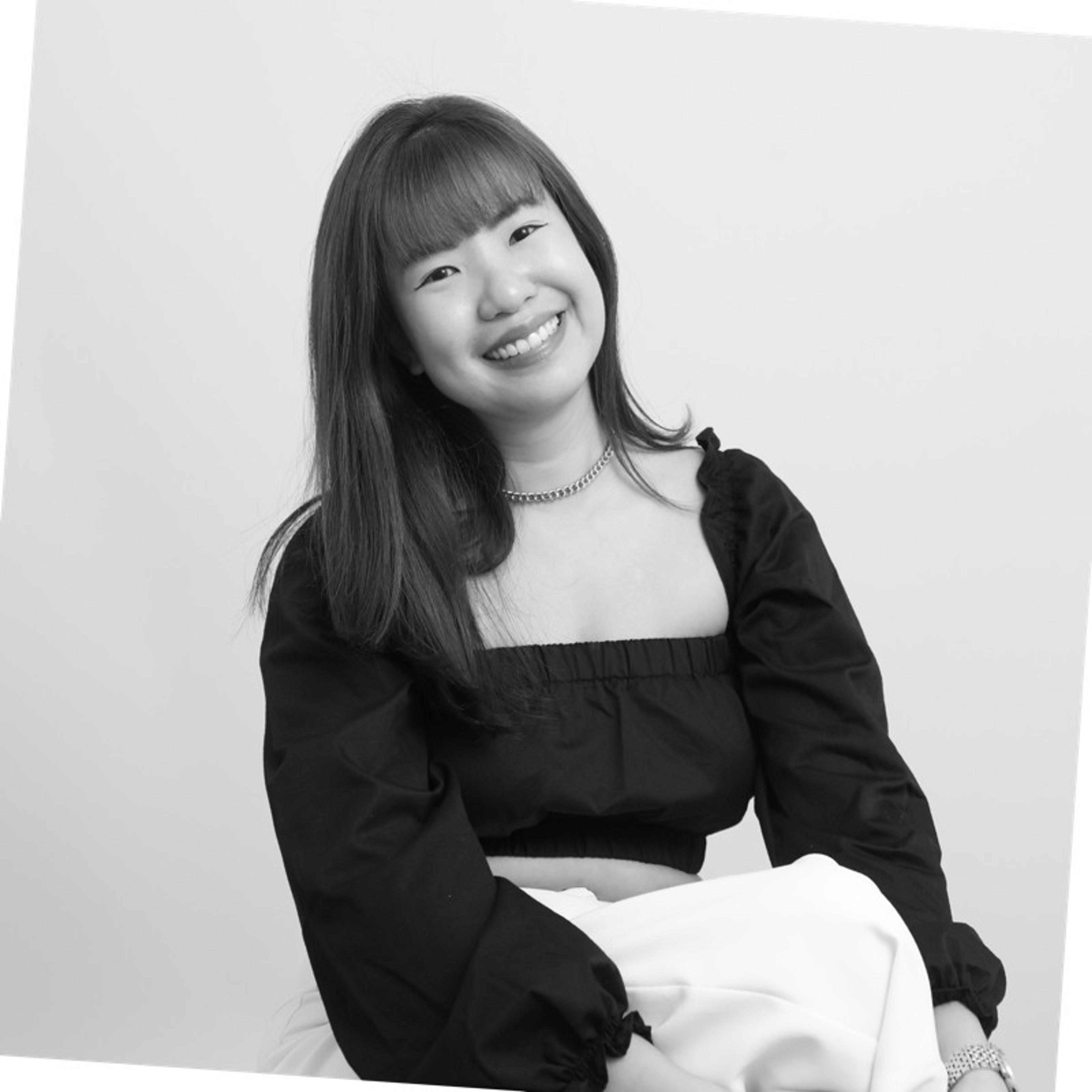 A smiling young woman with long hair sits against a light background, wearing a black off-the-shoulder top and a white skirt. She has a cheerful expression and is looking directly at the camera.