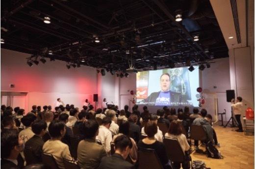 大勢の聴衆が集まる会場で、スクリーンに映し出された人物が話しています。参加者は講演を聞いています。