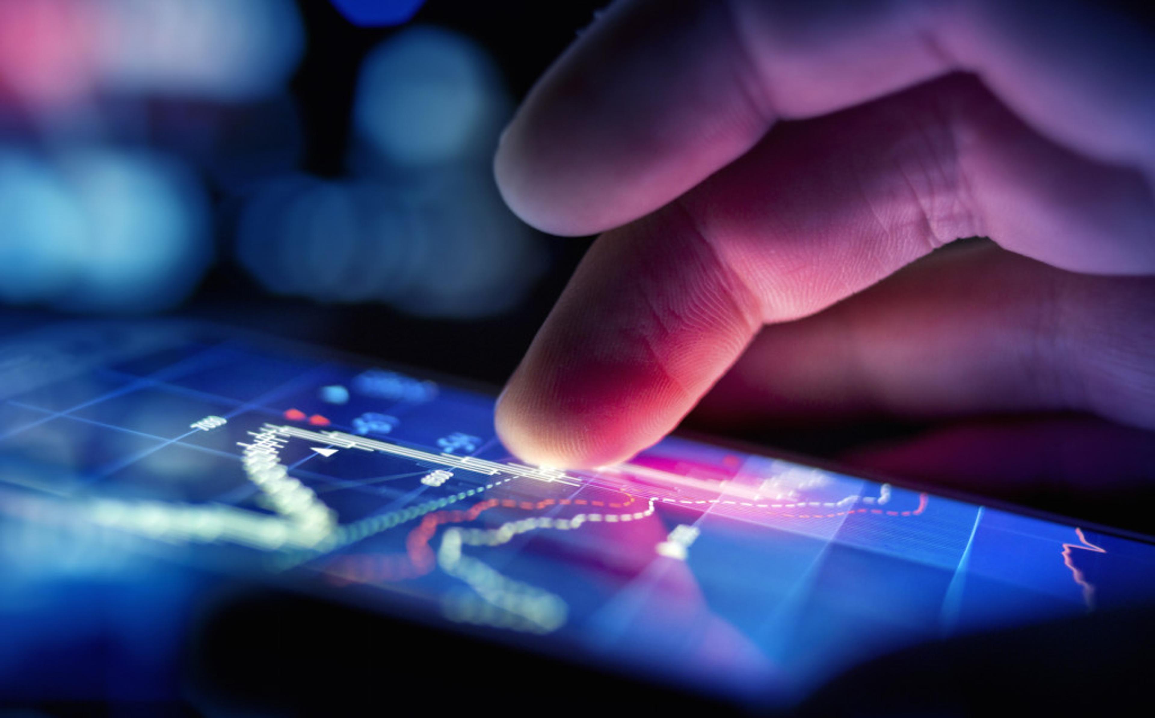 A close-up of a finger tapping on a smartphone screen displaying colorful financial graphs and data. The background is blurred with blue and purple lights.