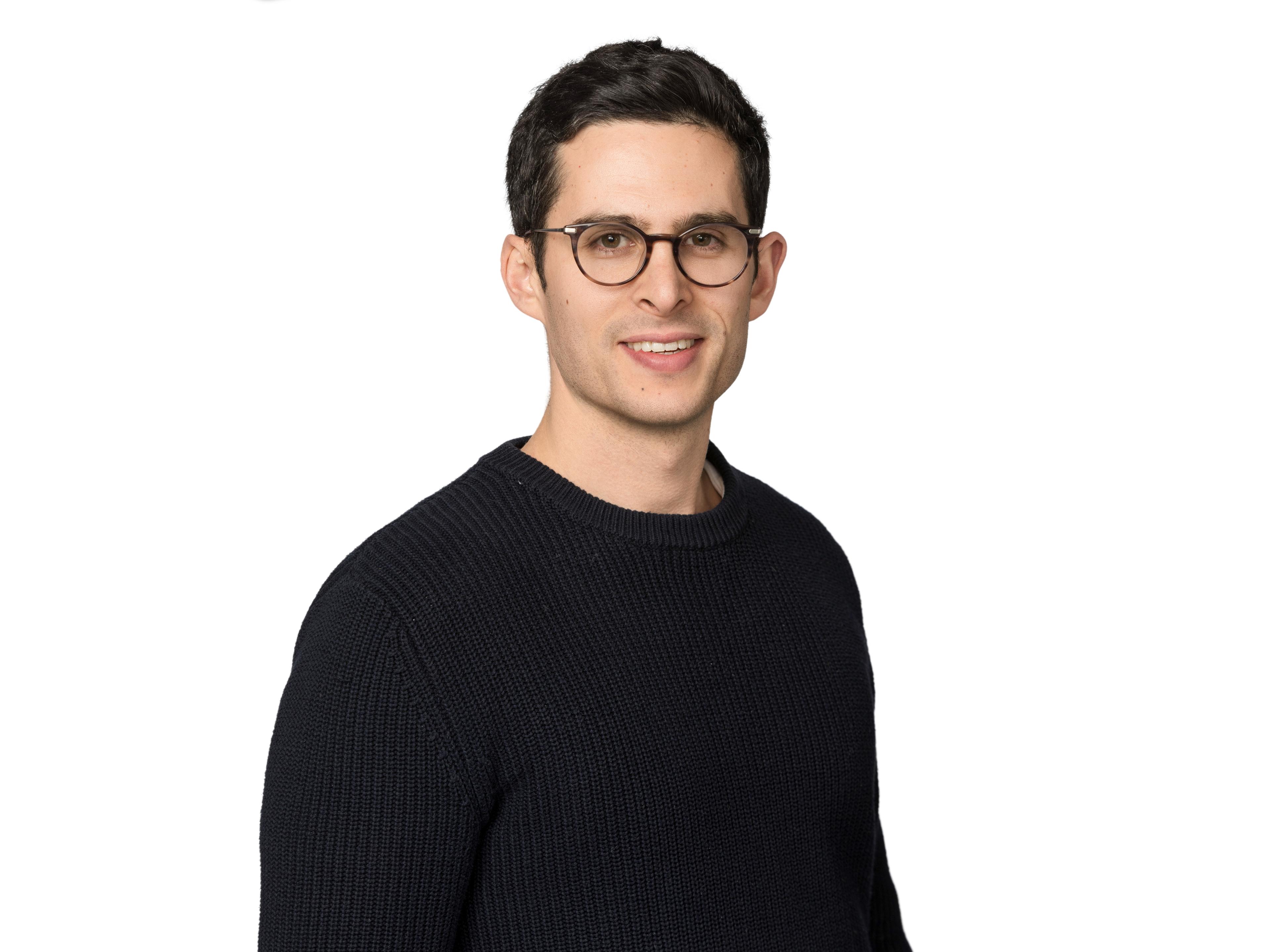 Hombre joven con gafas y suéter negro, sonriendo frente a un fondo blanco.