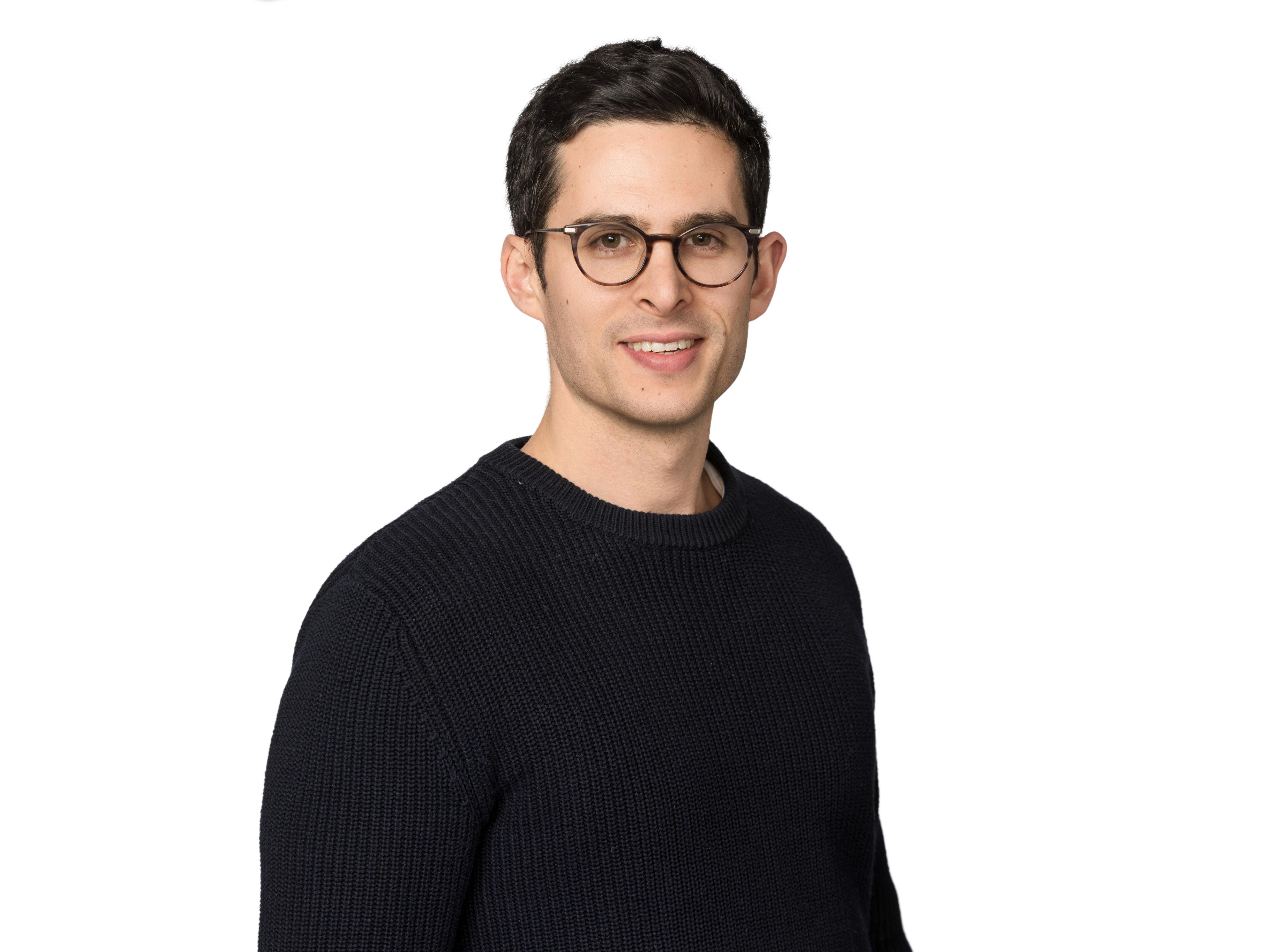 Hombre joven con gafas y suéter negro, sonriendo frente a un fondo blanco.