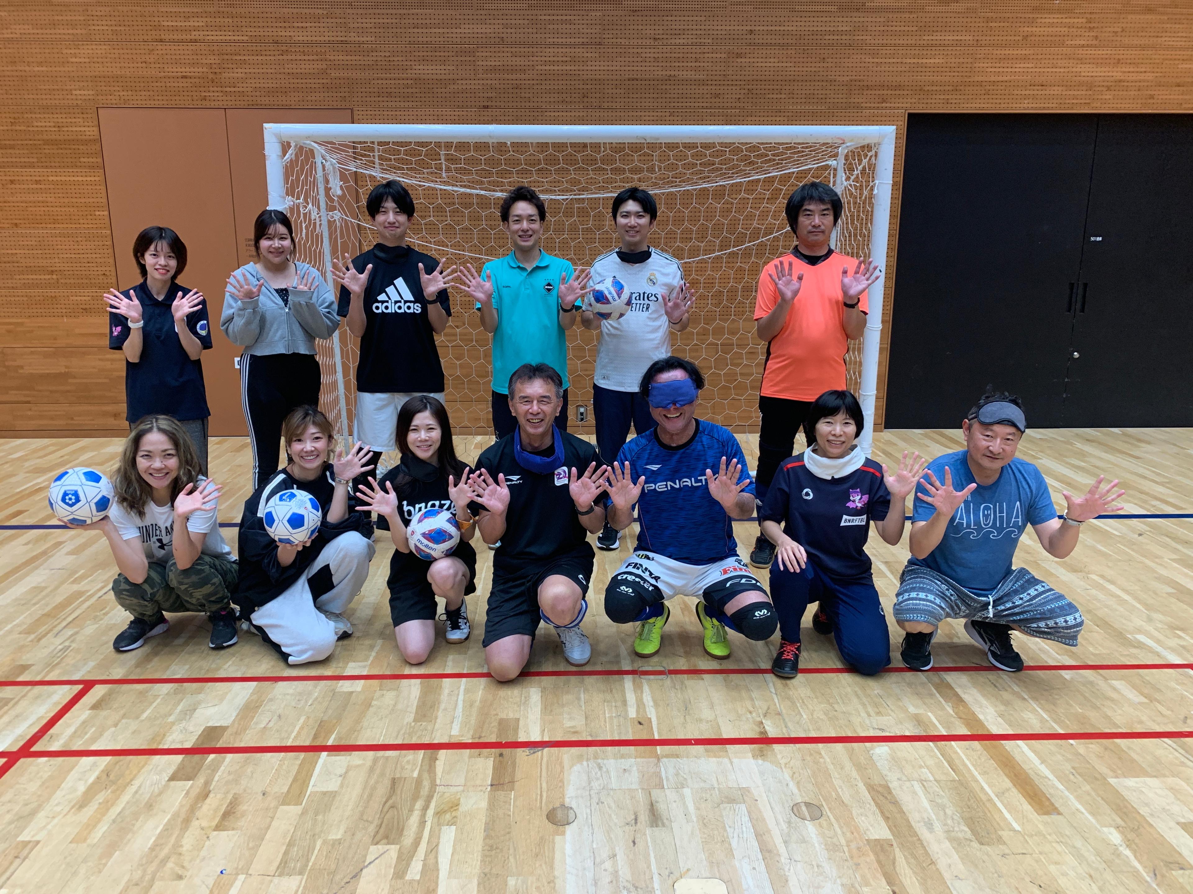 サッカーゴールの前でポーズをとるグループの人々。ボールを持った人や、手を挙げている人がいます。