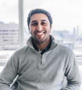 A smiling man with dark hair wearing a gray sweater sits in front of a window with a cityscape in the background.