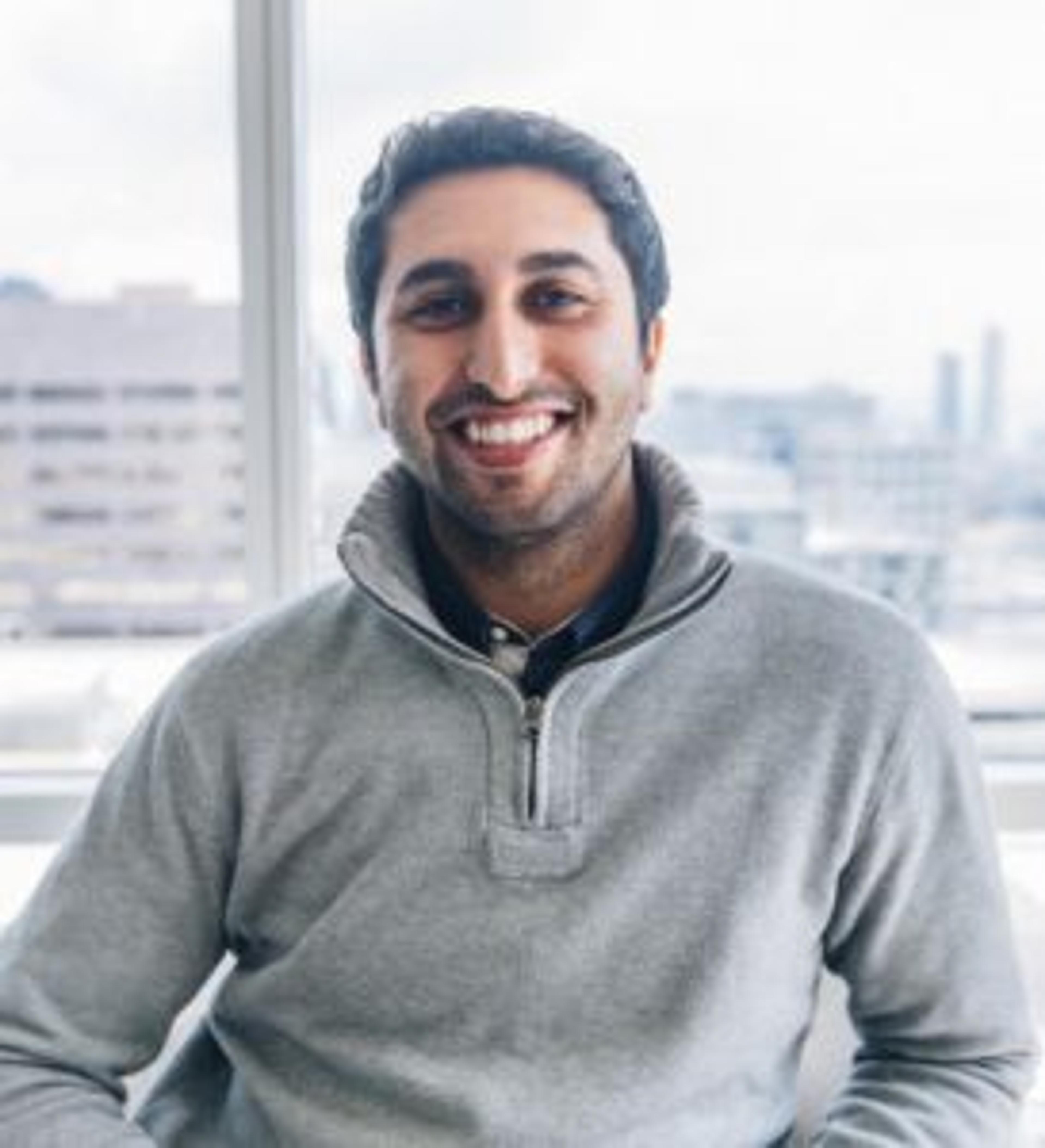 A smiling man with dark hair wearing a gray sweater sits in front of a window with a cityscape in the background.