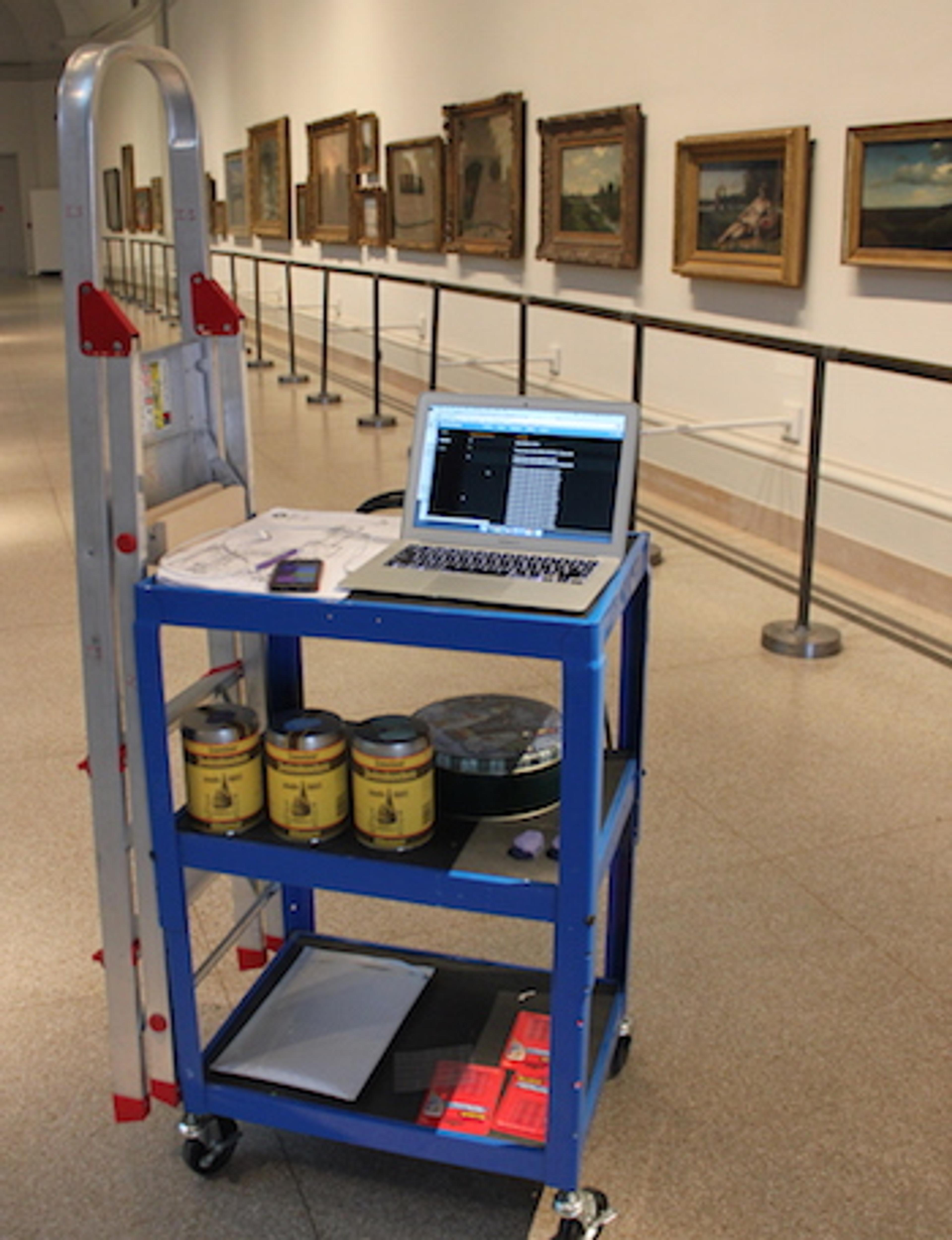 A blue cart with a laptop, paint cans, and supplies is positioned in an art gallery, with framed paintings displayed on the walls in the background. A ladder leans against the cart.