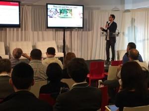 A speaker presents on stage to an audience seated in a conference setting. Two screens display presentation slides in the background.