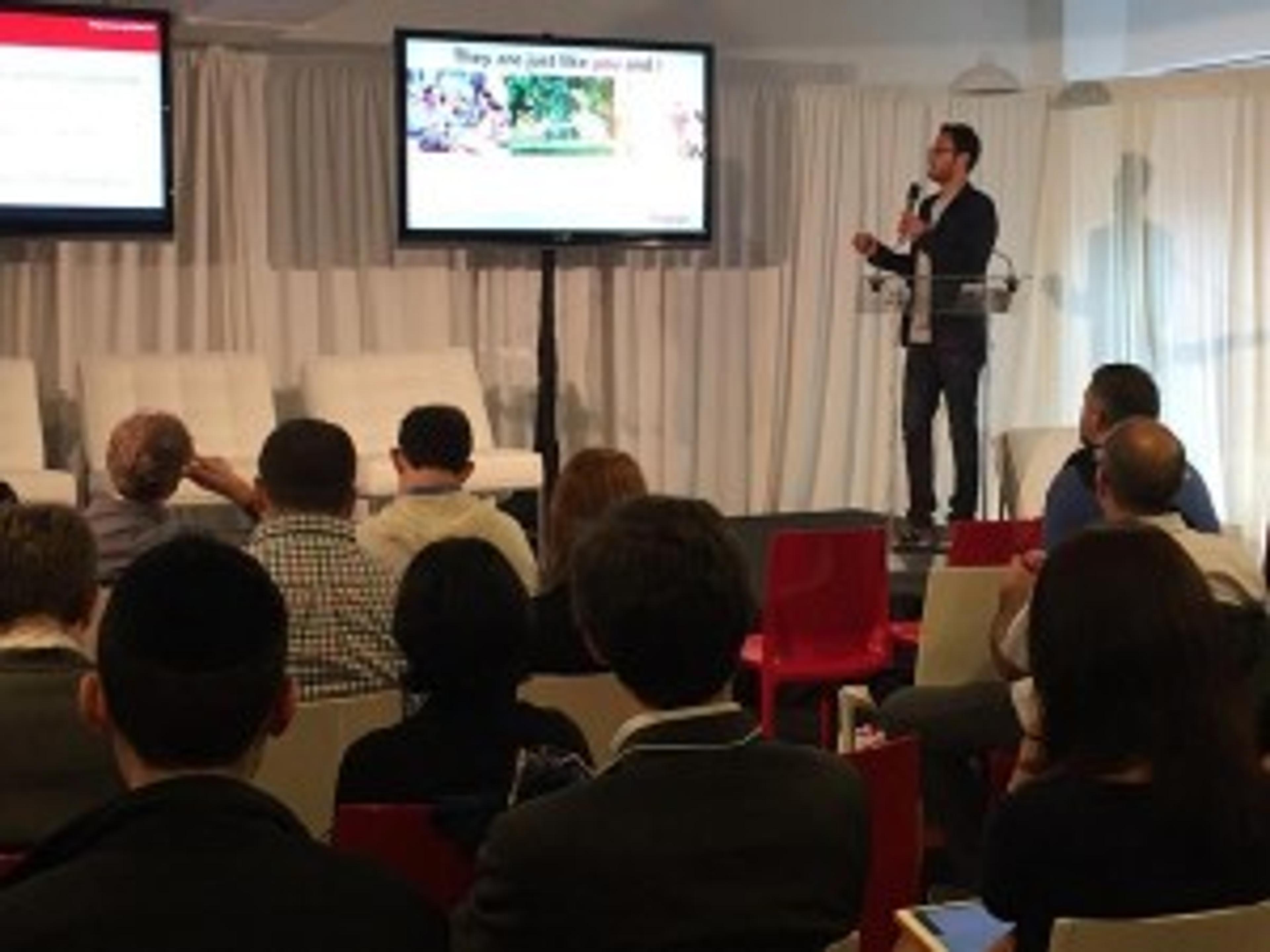 A speaker presents on stage to an audience seated in a conference setting. Two screens display presentation slides in the background.