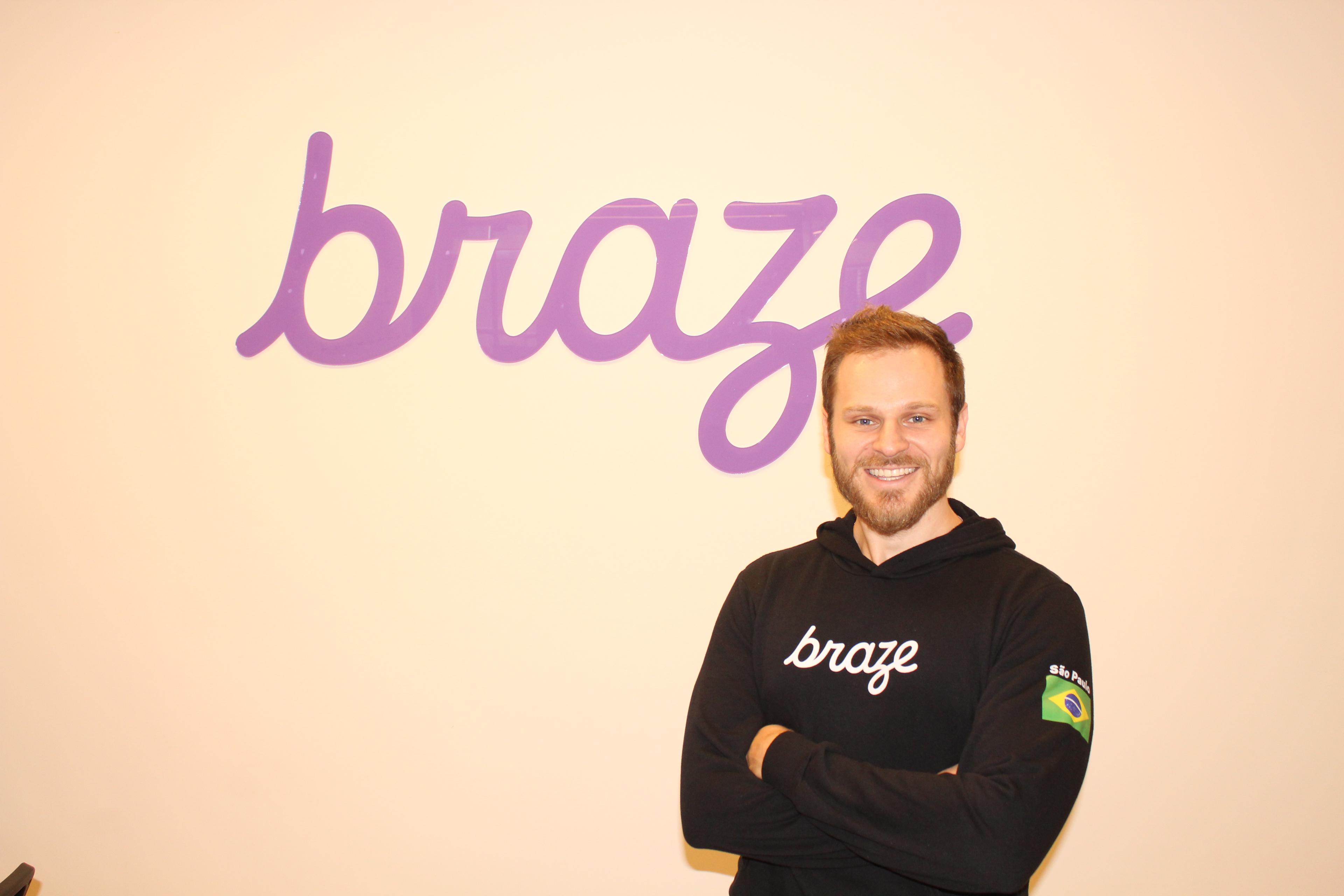 Hombre sonriente de pie frente a un logo de Braze en la pared. Lleva una sudadera negra con el logo de la empresa.