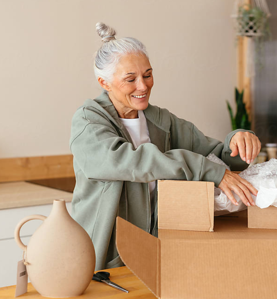 Smiling older woman in a casual jacket unpacks a cardboard box with bubble wrap on a kitchen counter, with a ceramic vase beside her.