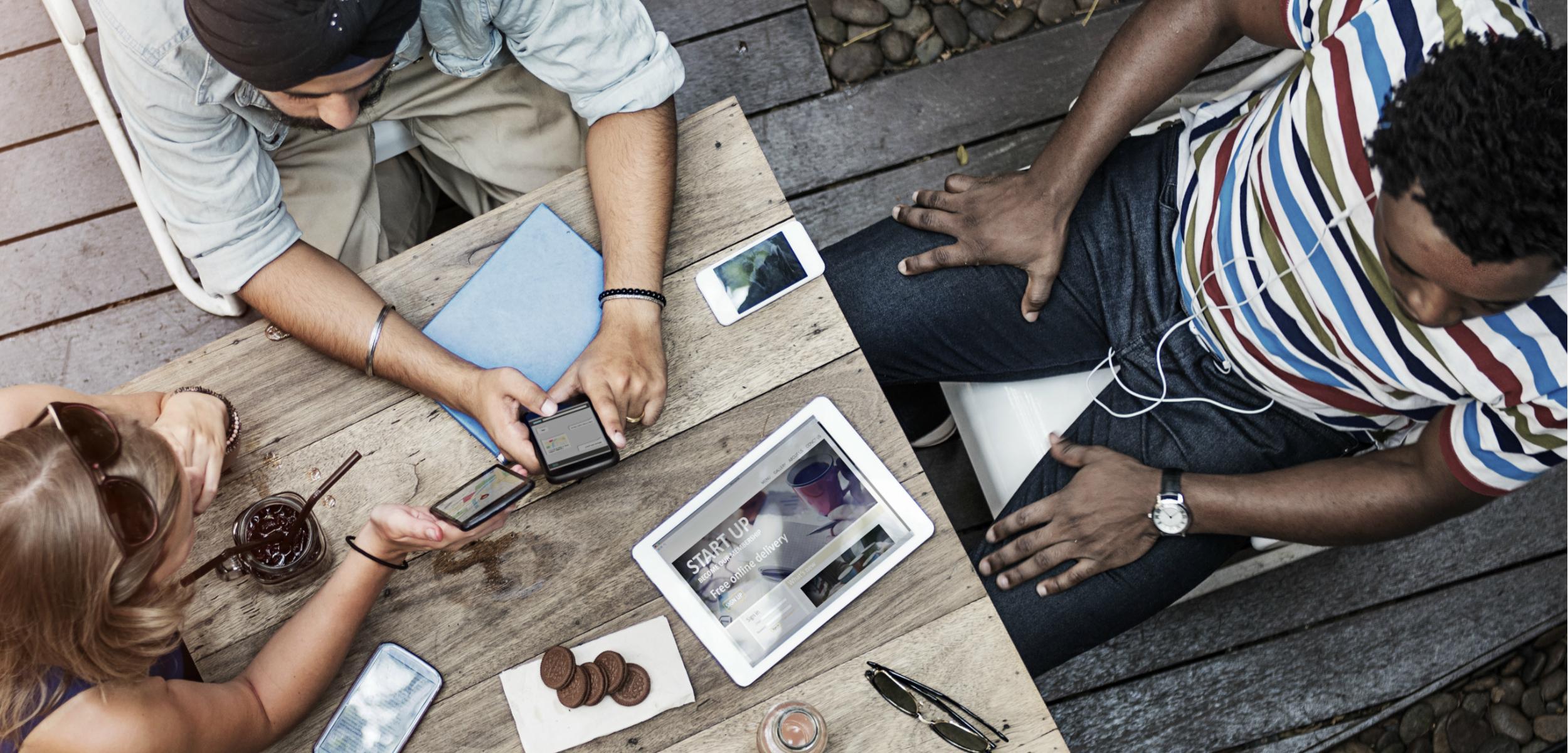 A group of three people sitting at a wooden table, using mobile devices and a tablet. There are drinks and snacks on the table.